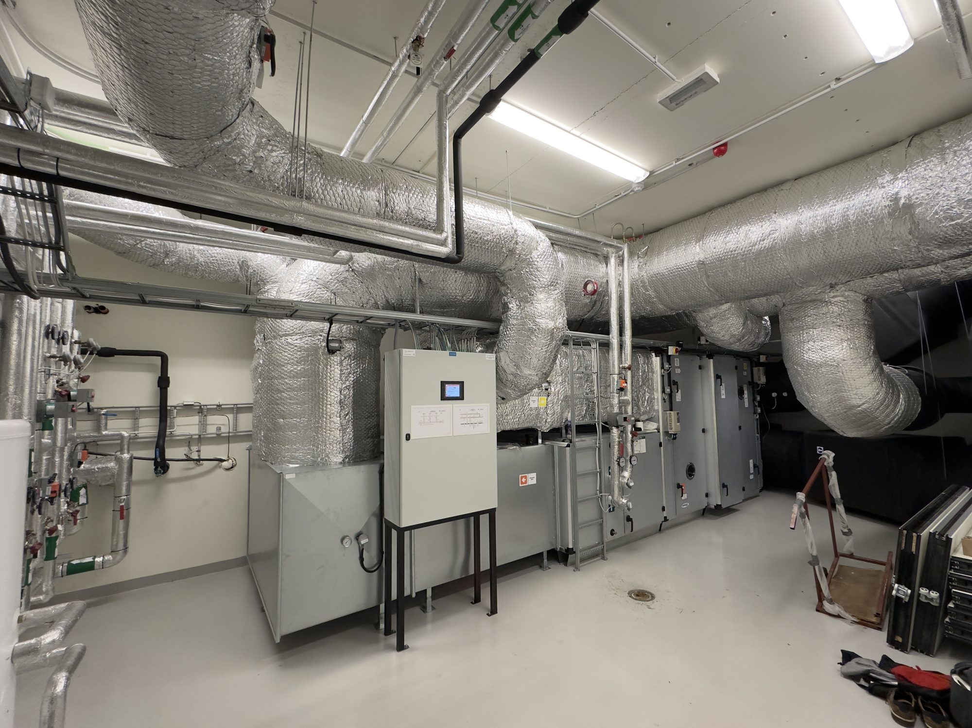Machines and equipment in the mechanical room (machine room) of a school building in Lillehammer, Norway, with a generator for the heating system, ventilation and air conditioning. The room contains the following, among other things:
Main distributors for the heating system and air handling, ventilation circuits, radiator circuits, underfloor heating circuits
OSO water heater with a tank for hot water
Insulated pipes with thermometers (measuring instruments, gauges with round displays and needles), circulation pump, throttle valve, radiators, underfloor heating
Switches, controls
Water tap on the wall
Heat recovery unit (which reuses heat already present in the building), generator, ventilation, air, air filter, treated supply air, exhaust air, exhaust fan
Inspection ladder
Electrical panels (distribution boards, circuit breakers) in a closed cabinet
Power cables (electrical wiring) mounted on the wall and ceiling tracks, etc.
Storage of equipment
Utility sink with faucets
Fire extinguisher
etc.
Photographs (mobile phone pictures) of the room and details were taken in September 2024, some with a wide-angle lens