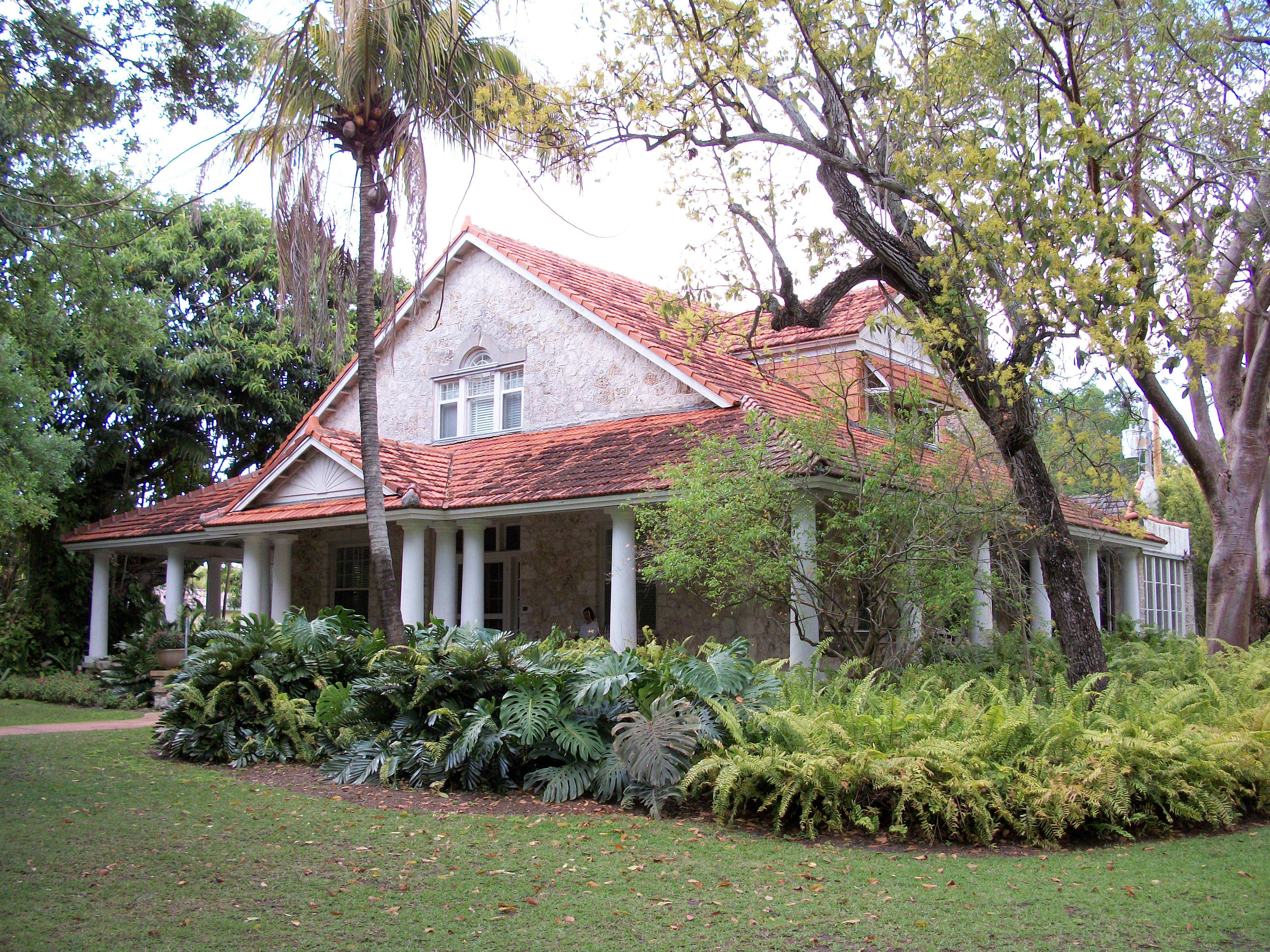 Historic house with white columned wraparound porch surrounded by tropical vegetation.