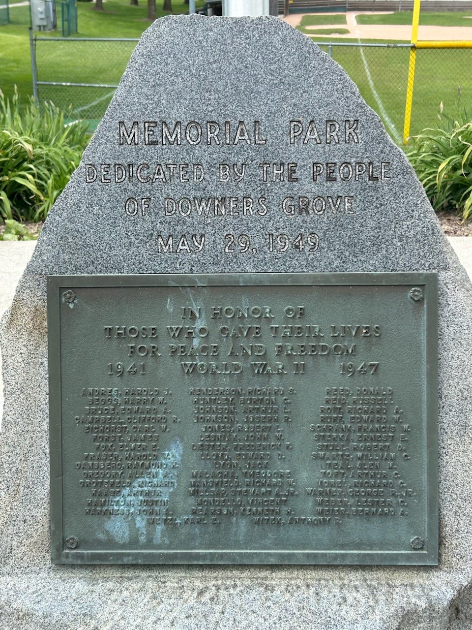 Granite memorial stone with bronze World War II plaque in a park setting.