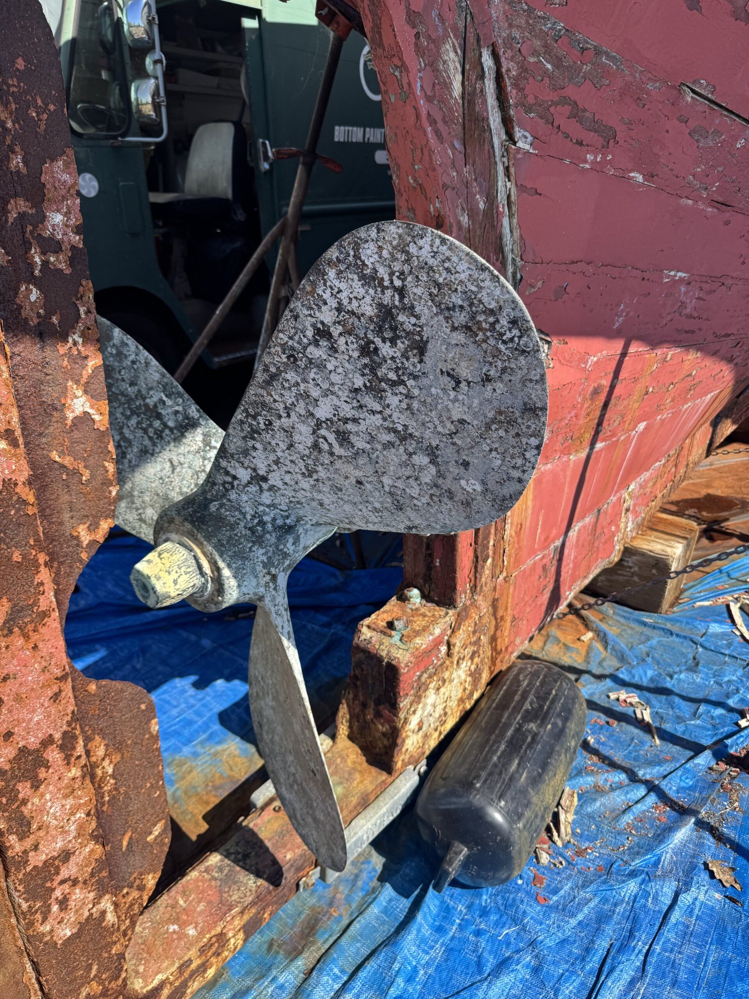 Weathered metal boat propeller mounted on a stand with a rusted red hull in the background.