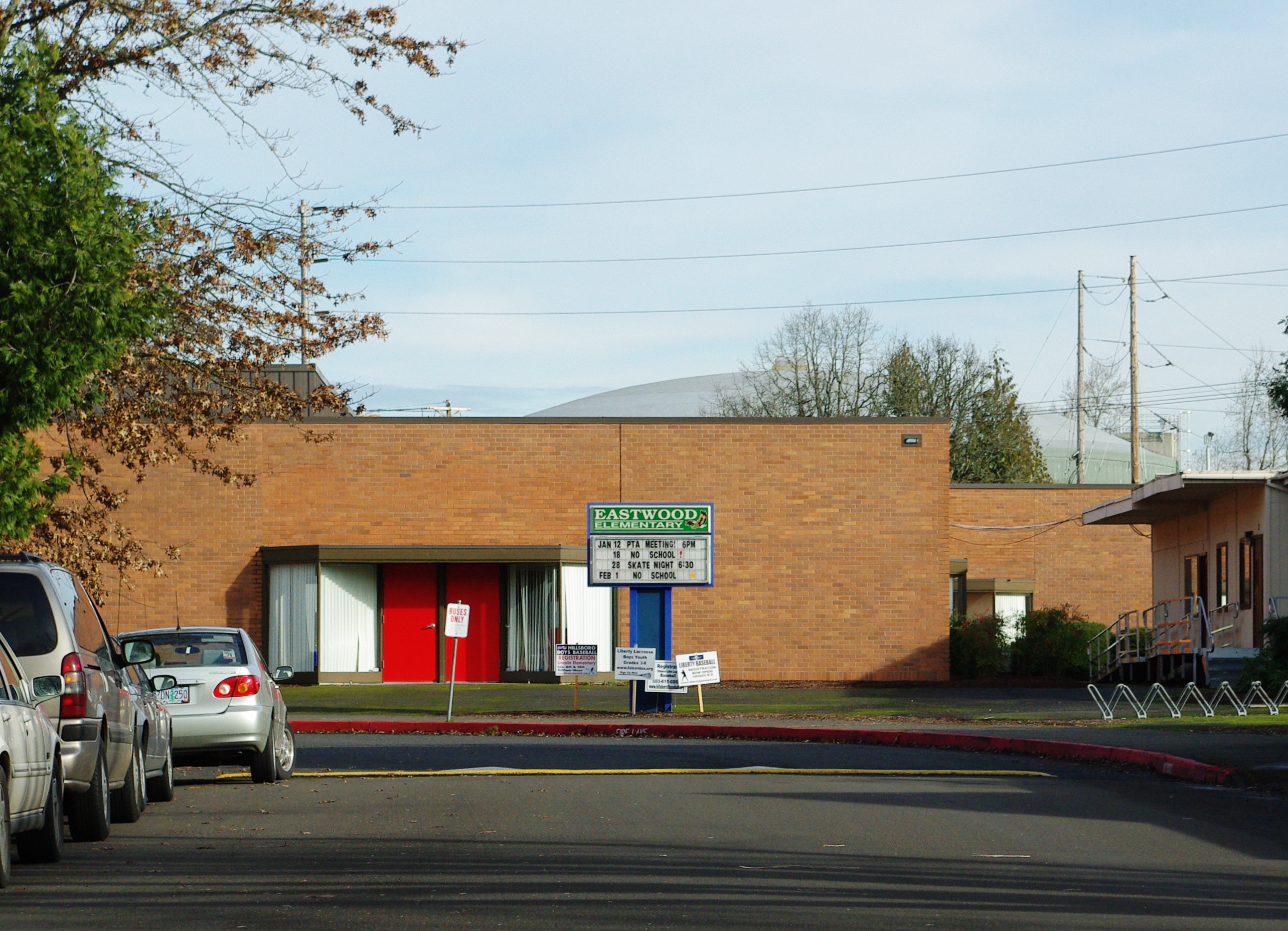Brick elementary school building with red entry doors and a marquee sign on a tree-lined street.