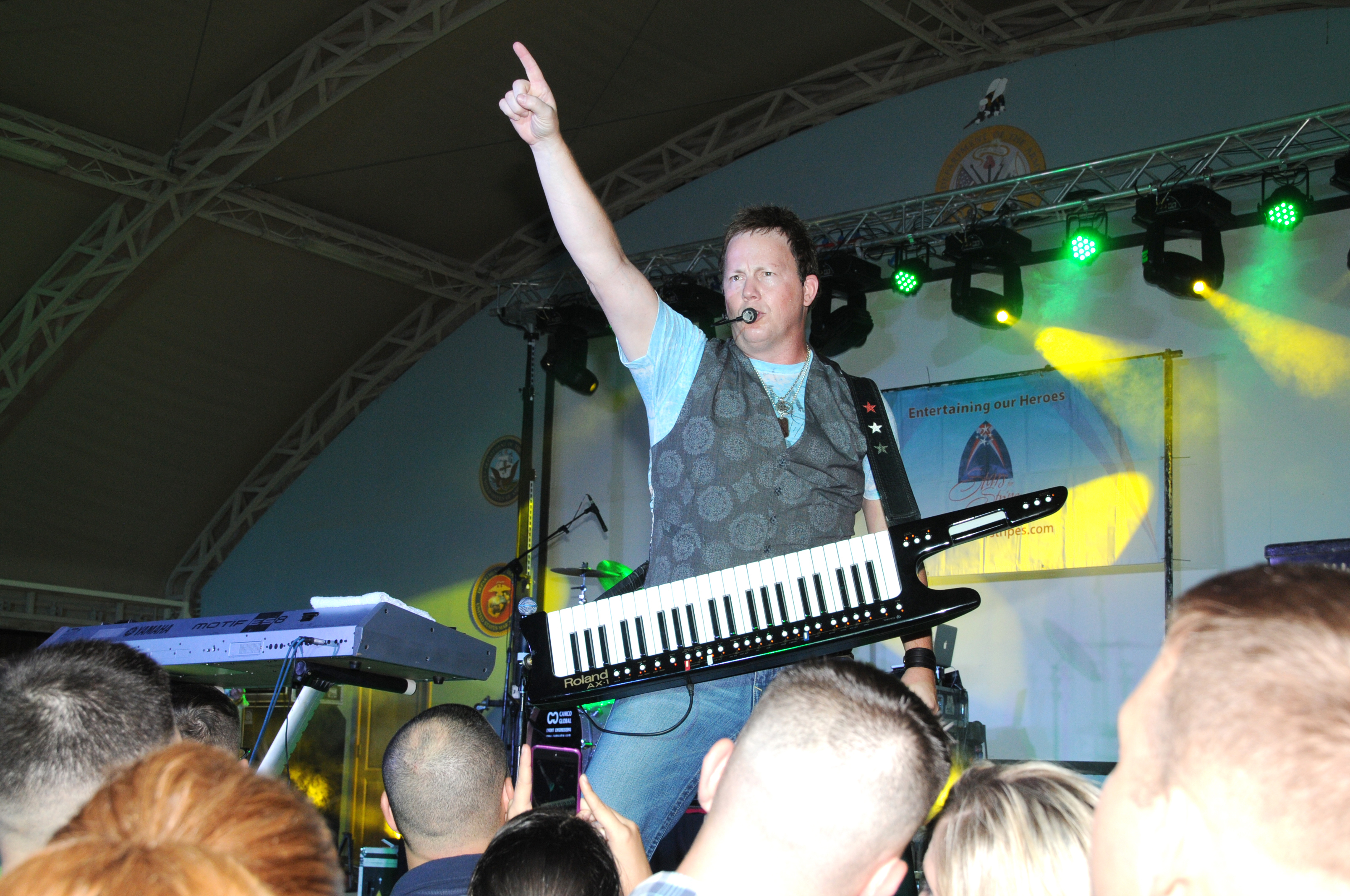 A performer on stage plays a keytar with one arm raised, crowd in foreground.
