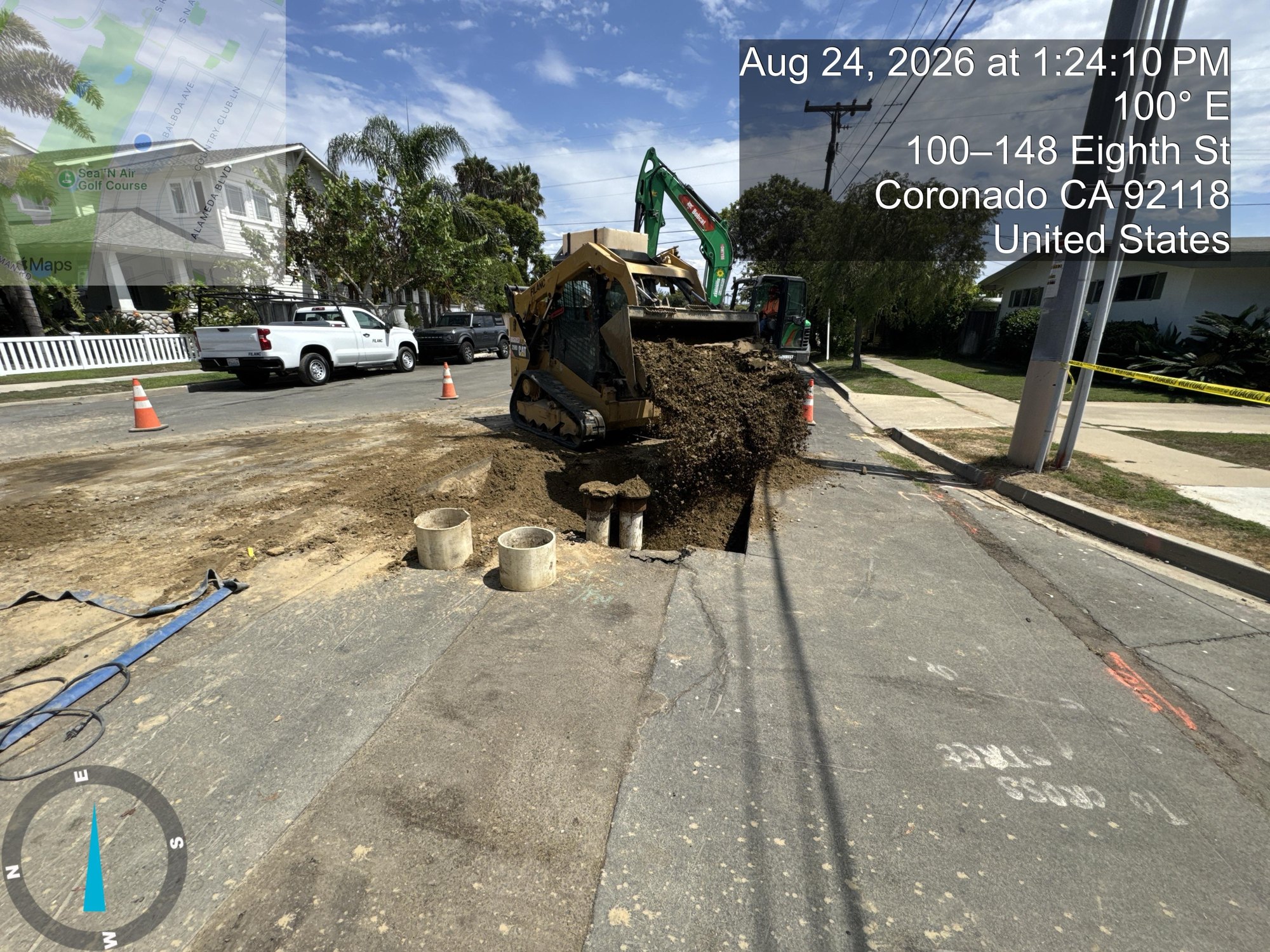 Green excavator digging a trench with large concrete pipes on a residential street.