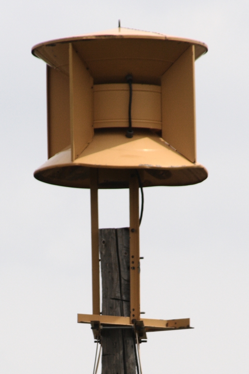 Tan civil defense outdoor warning siren on a wooden utility pole against a gray sky.