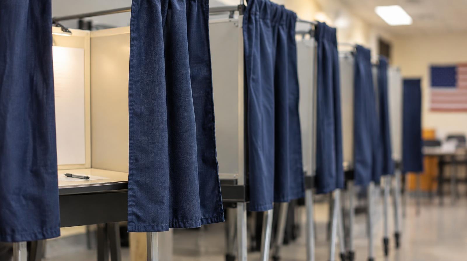 A row of empty voting booths with privacy curtains under fluorescent lighting.