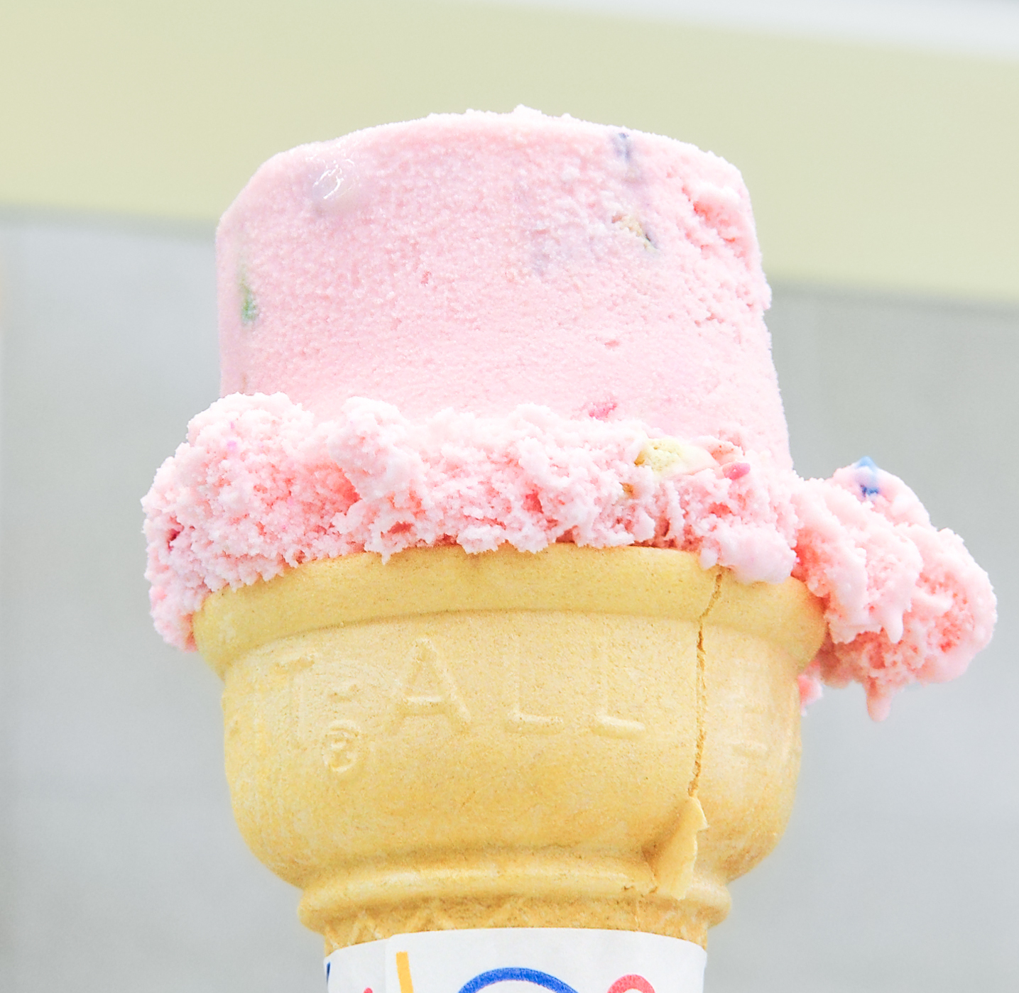 A single scoop of pink ice cream melting over the rim of a cake cone.