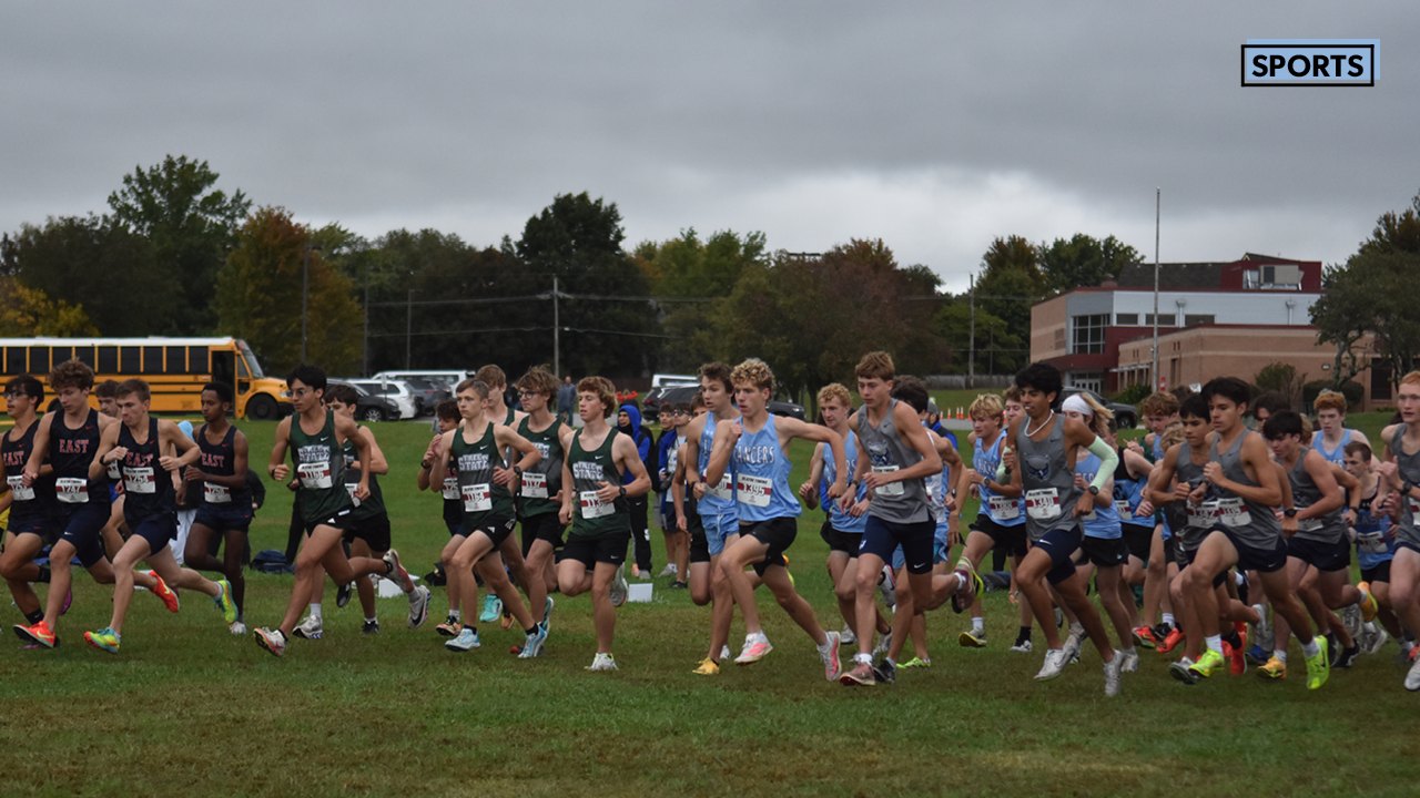 A large group of runners in numbered bibs starts a cross country race on a grass field.