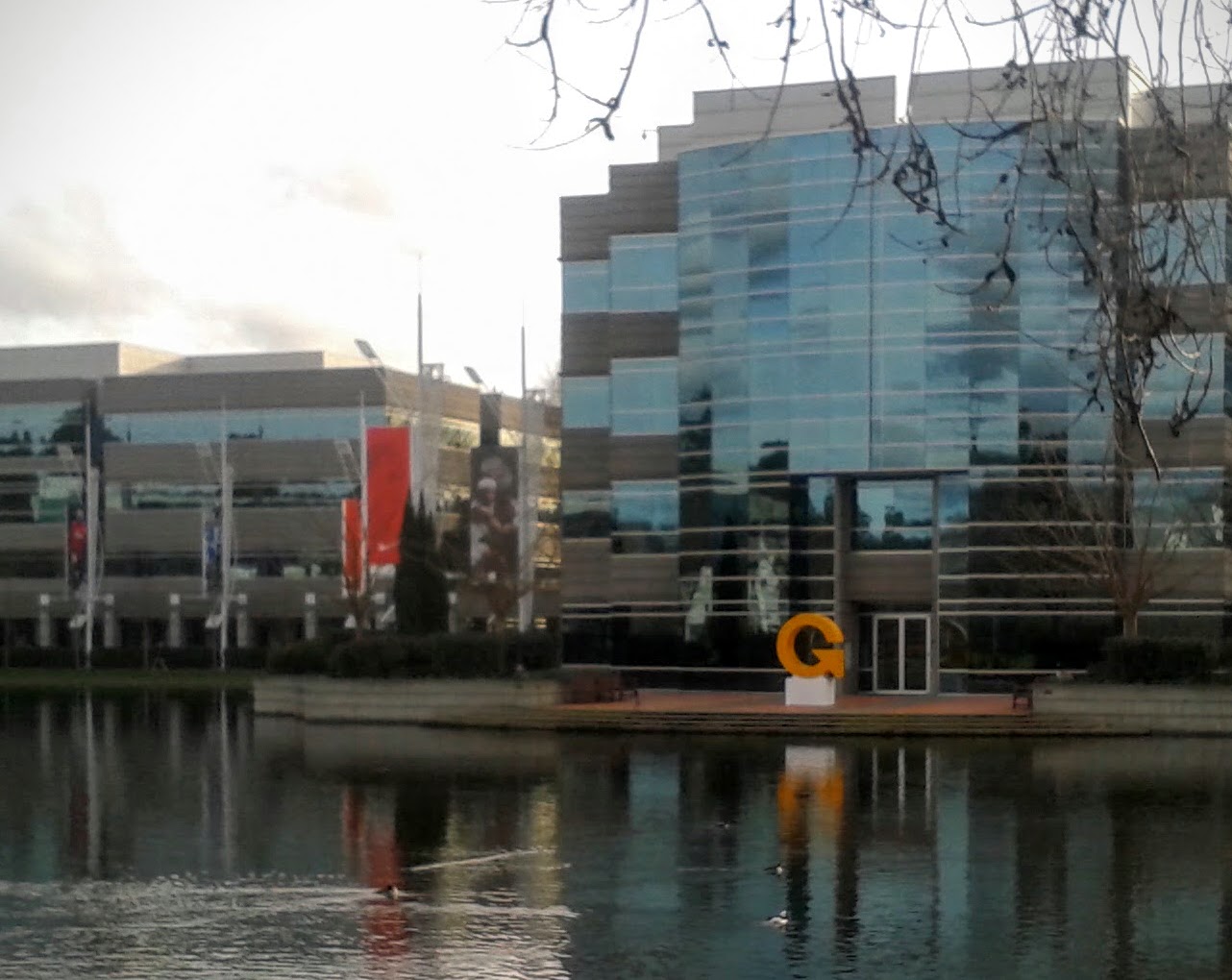 Glass office building at a corporate campus reflected in a pond, with a sculpture on a dock in the foreground.