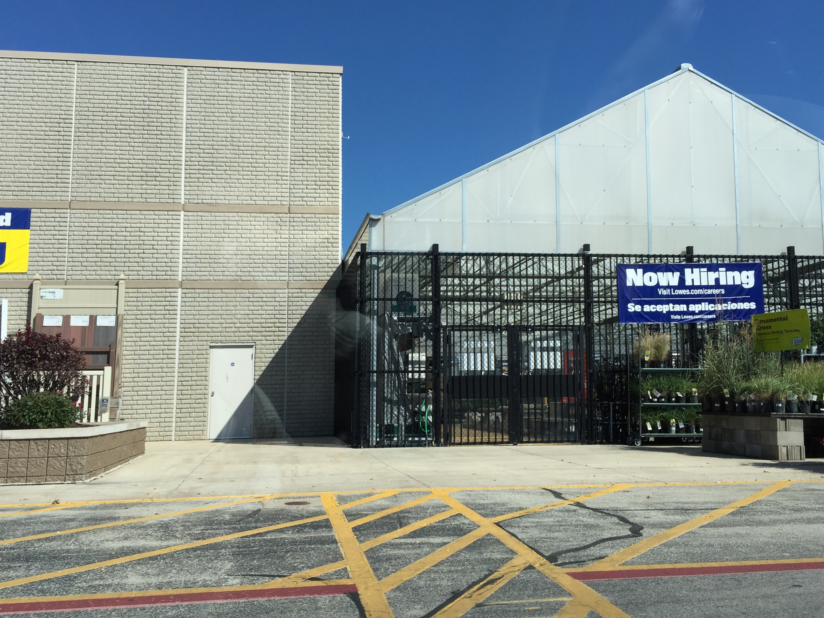 Exterior of a Lowe's garden center with iron gates, greenhouse structure, and a Now Hiring sign.