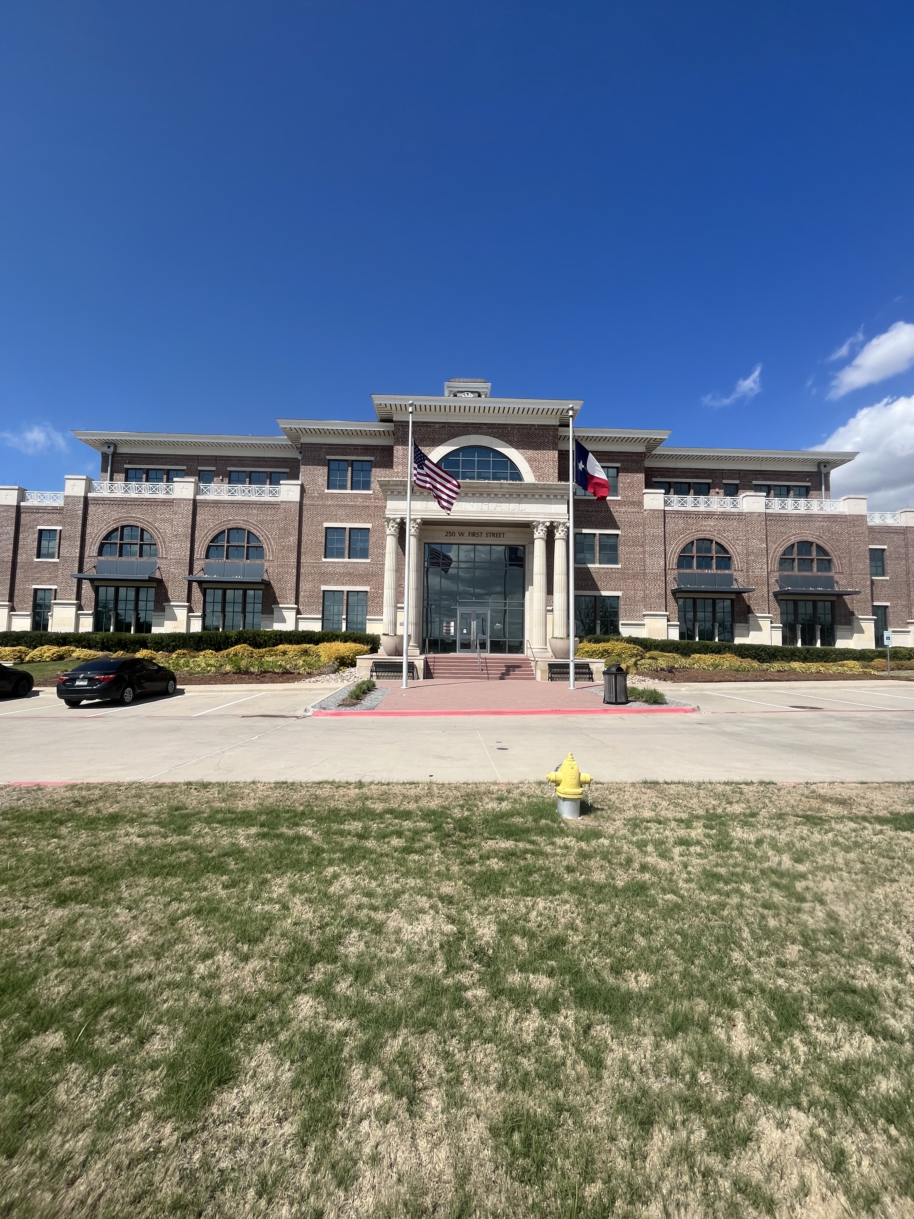 Two-story brick town hall with arched windows and U.S. and Texas flags on front lawn.