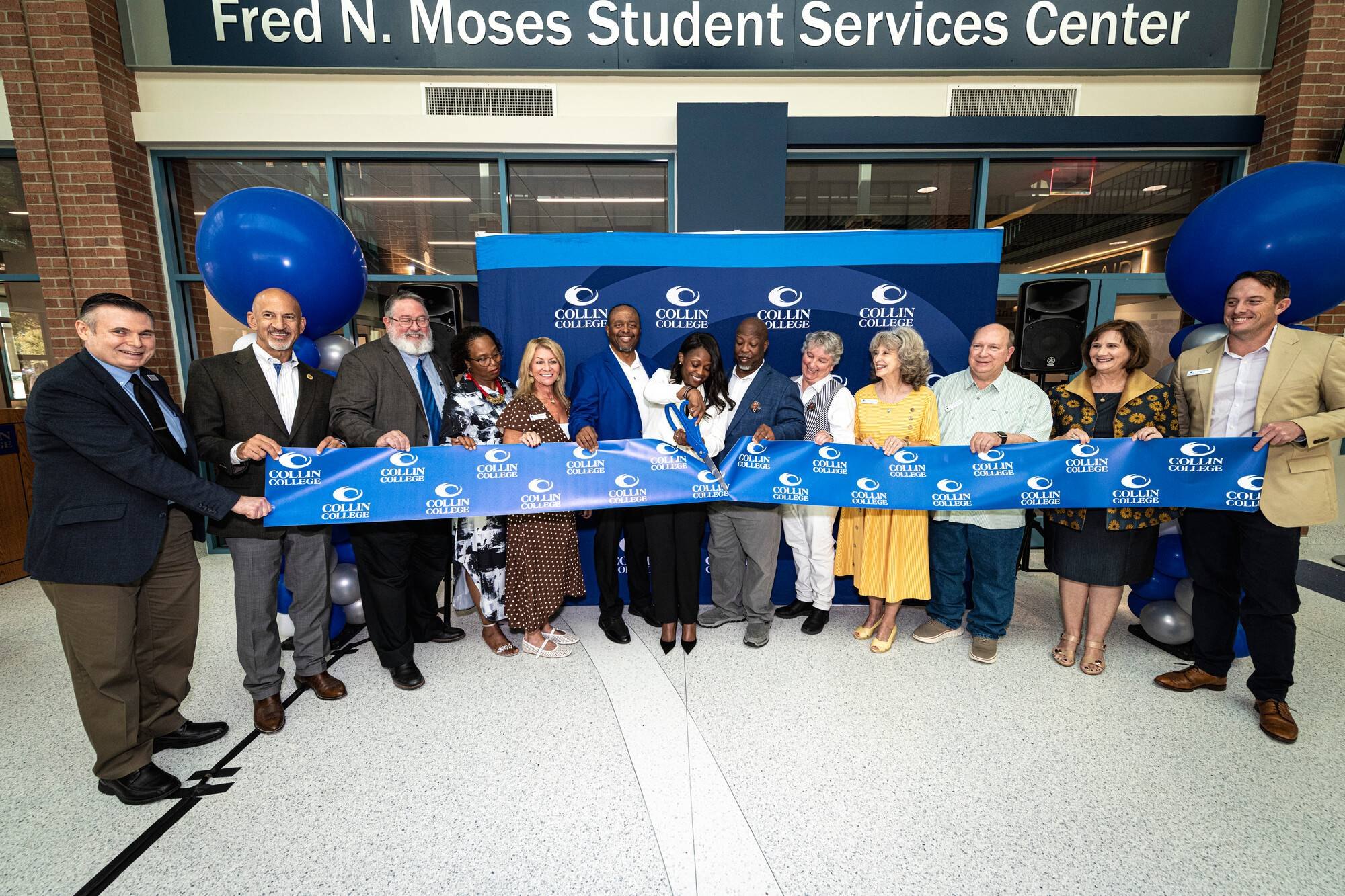 Group of people holding a blue ribbon in front of a building entrance with blue balloons.