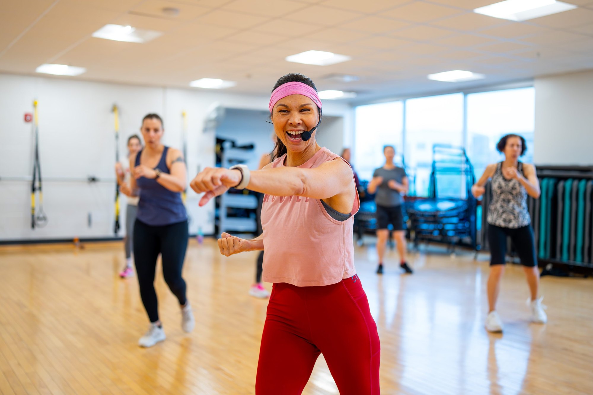 A woman in a pink top leads a group fitness class in a mirrored gym studio.