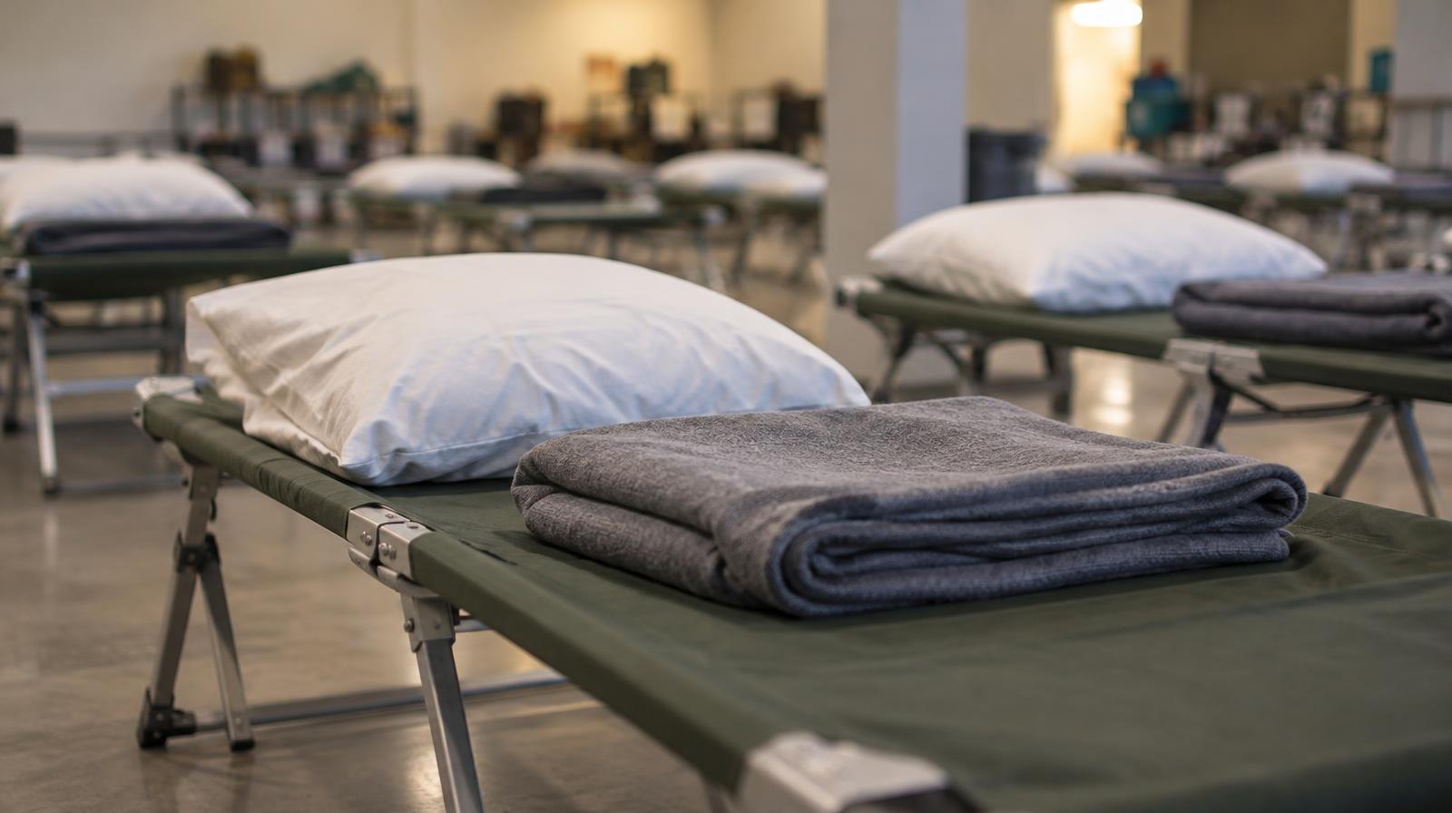 Rows of neatly made cots with folded blankets inside a shelter.