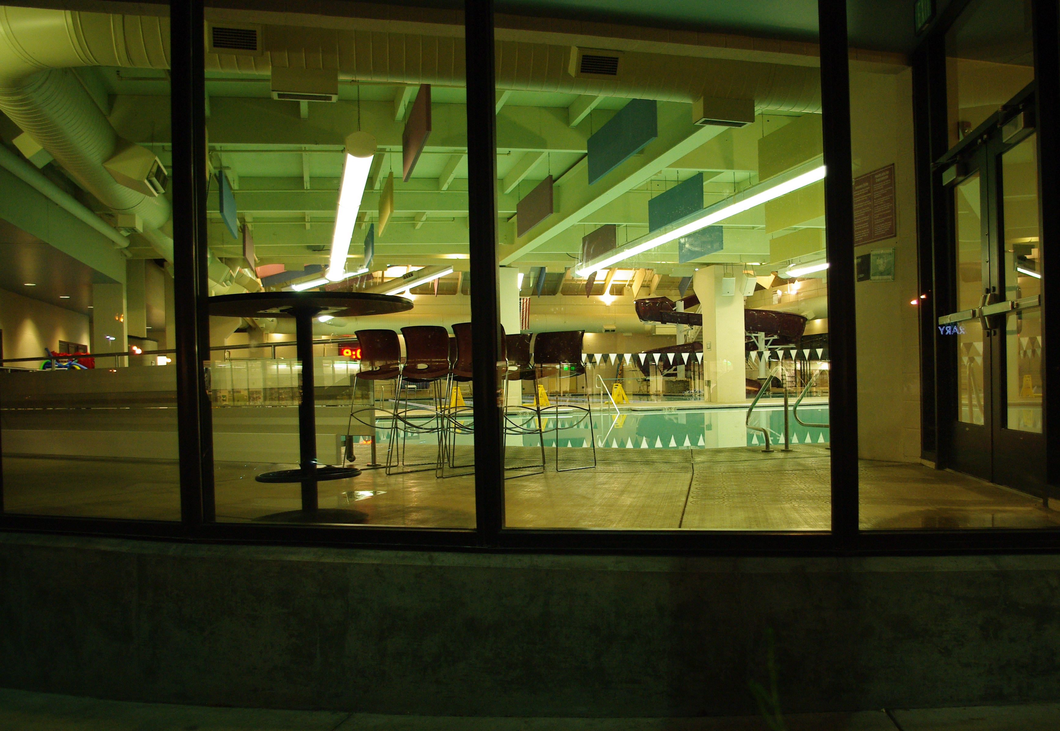 Indoor pool with lane dividers and overhead lights, viewed through large windows at night.