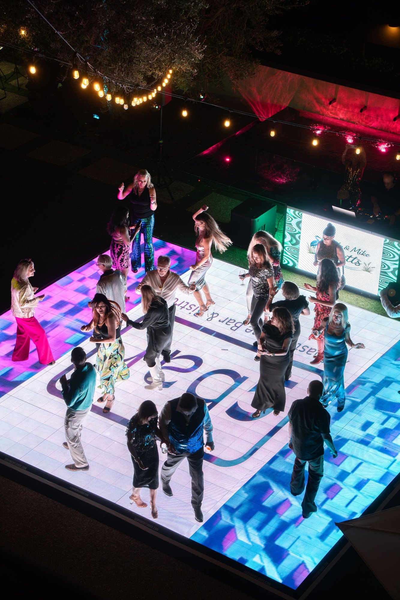 Guests in colorful sequined outfits dance on a large illuminated LED dance floor at an outdoor evening event.