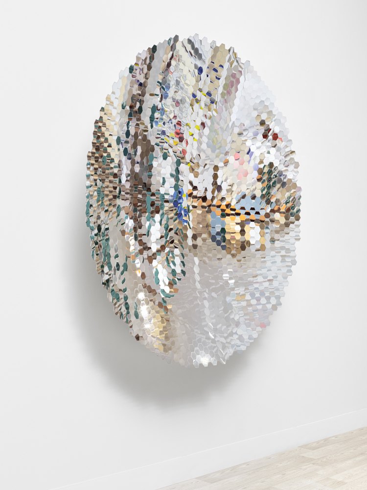 Large oval wall sculpture made of hexagonal mirrored tiles reflecting light in a gallery.
