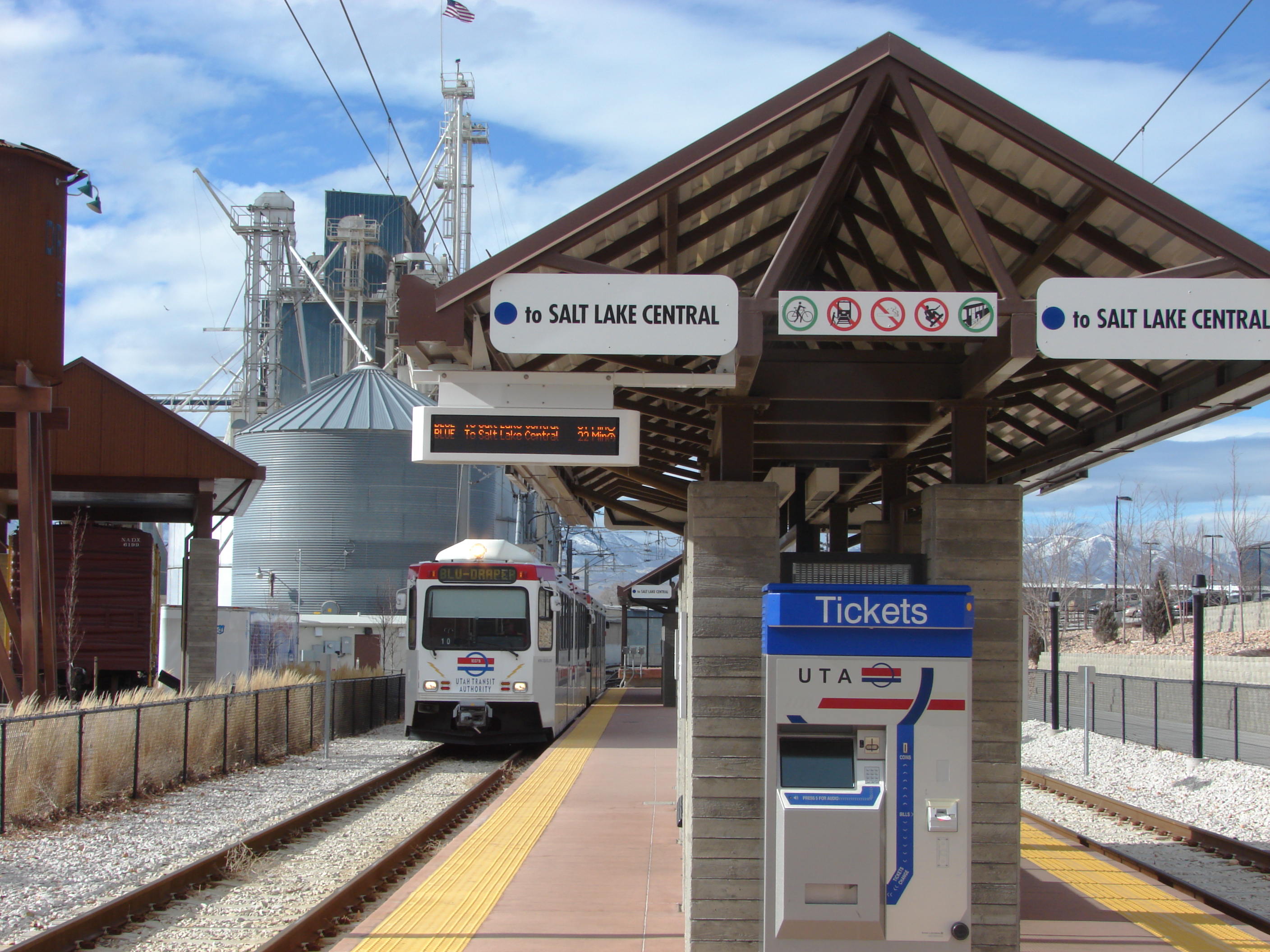A TRAX light rail train pulls into a covered station platform with directional signs overhead.