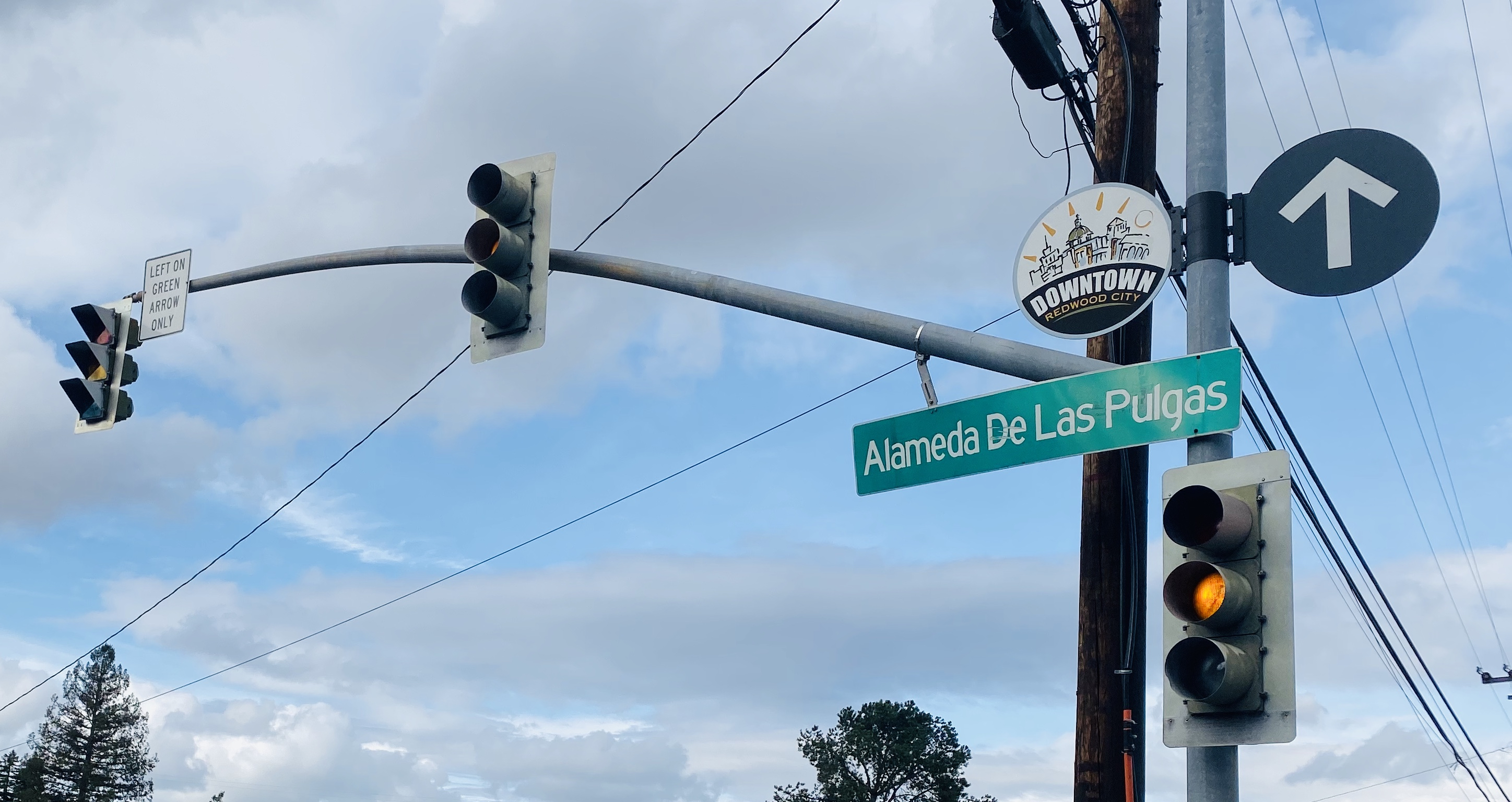 Green street sign for Alameda de las Pulgas on a pole at an intersection with traffic lights.