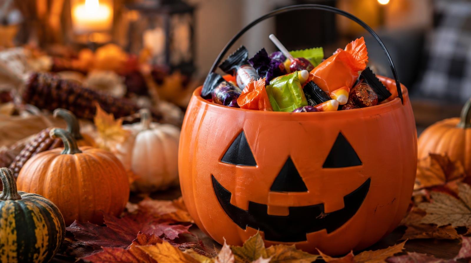A jack-o'-lantern bucket overflowing with Halloween candy surrounded by small pumpkins and autumn leaves.
