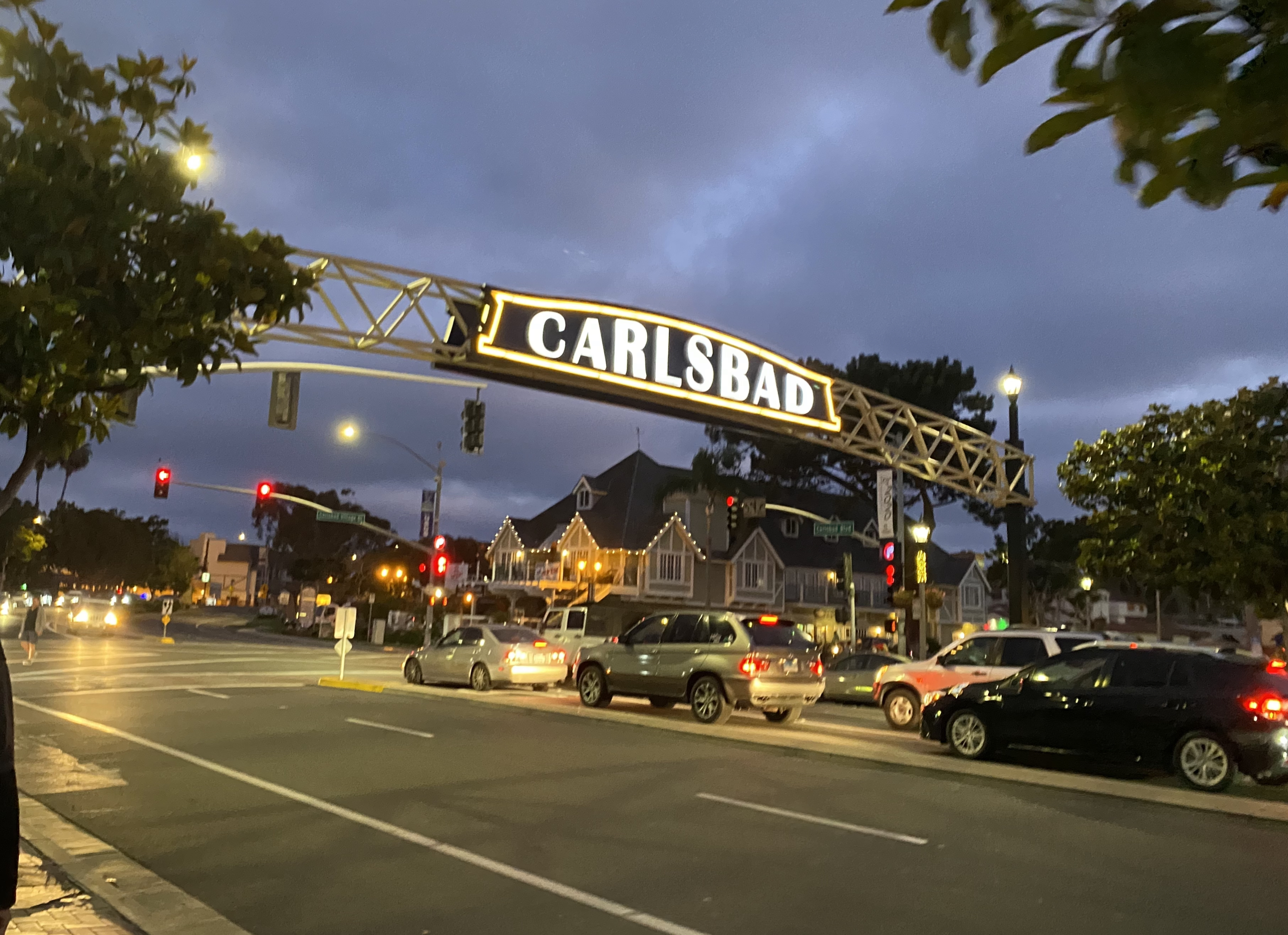 Lit arch sign reading CARLSBAD spans a boulevard at dusk with traffic below.