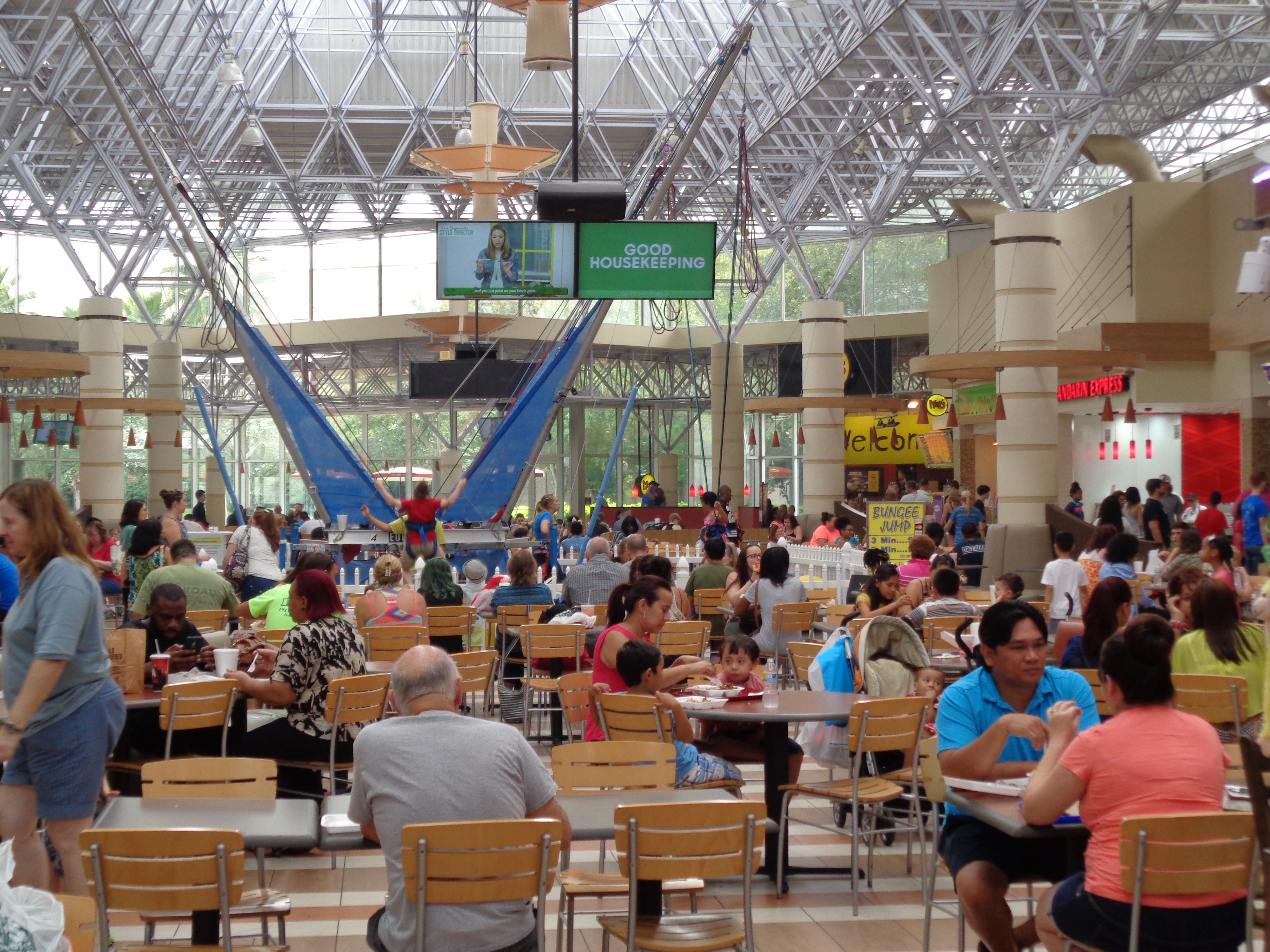 Crowded indoor mall food court with shoppers seated under a glass skylight ceiling.