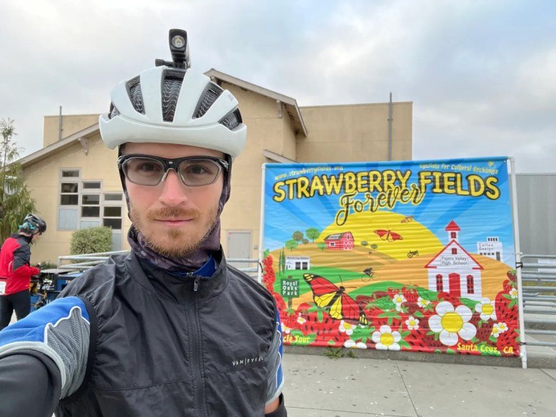 A man in a white cycling helmet and jacket takes a selfie in front of a colorful mural.