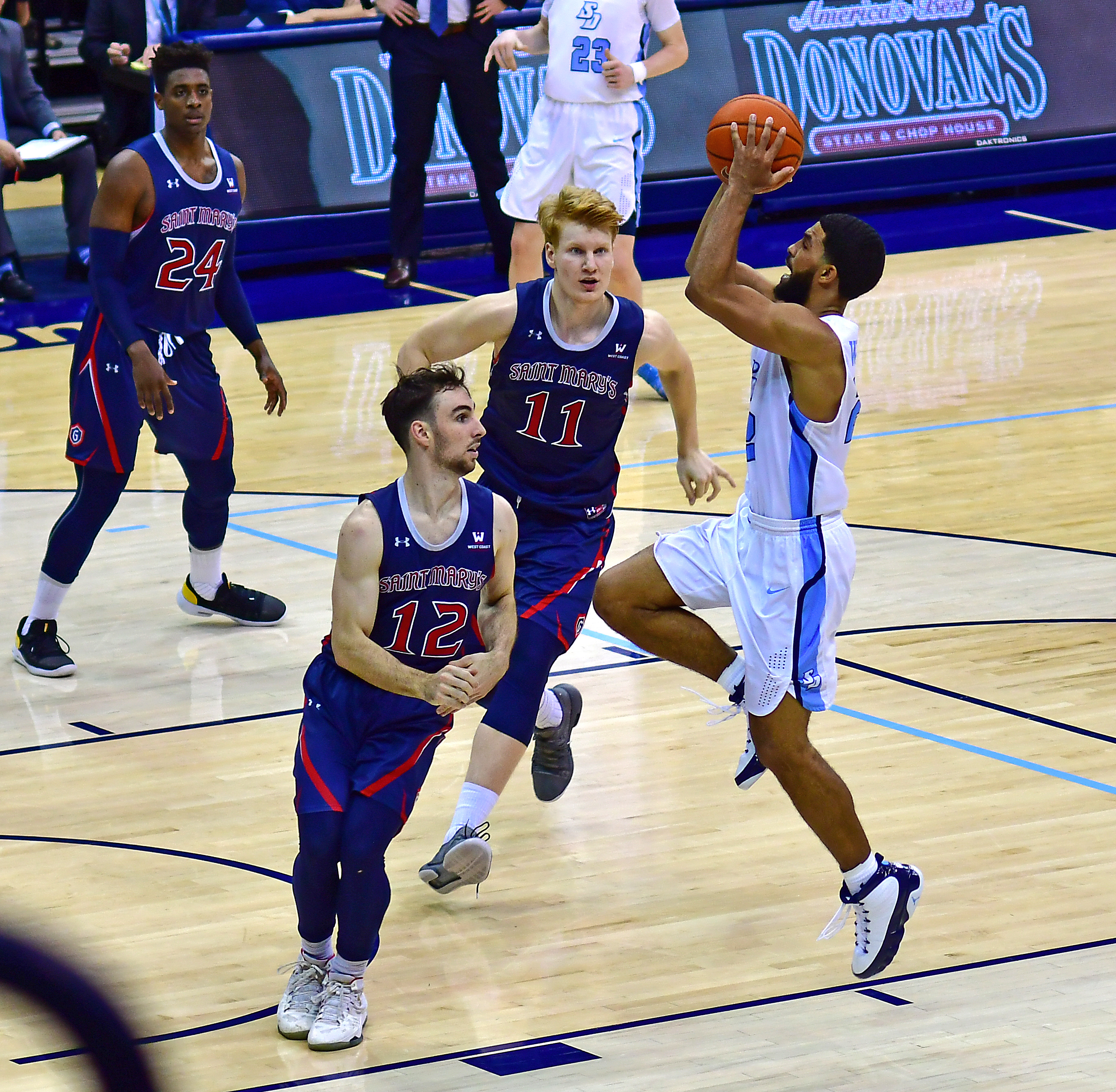 A basketball player in a white uniform leaps to shoot over two defenders.