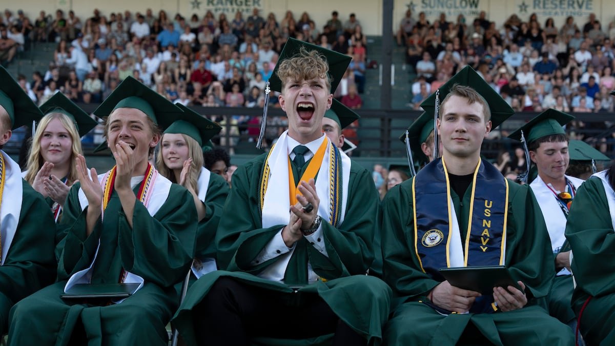 Graduates in green caps and gowns clapping and cheering at an outdoor graduation ceremony.