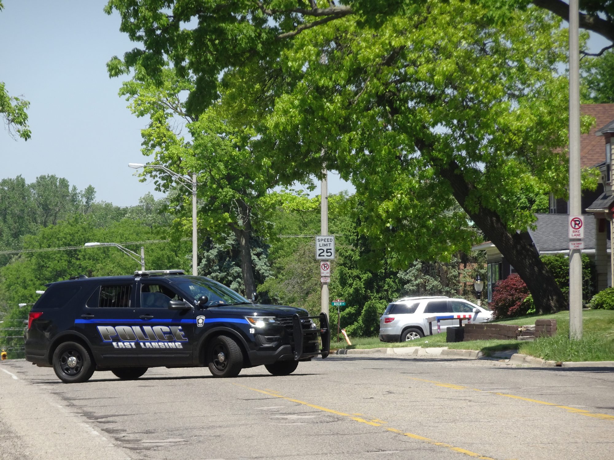 An East Lansing Police Department SUV blocks a tree-lined residential street on a sunny day, with a parked car and houses visible in the background.