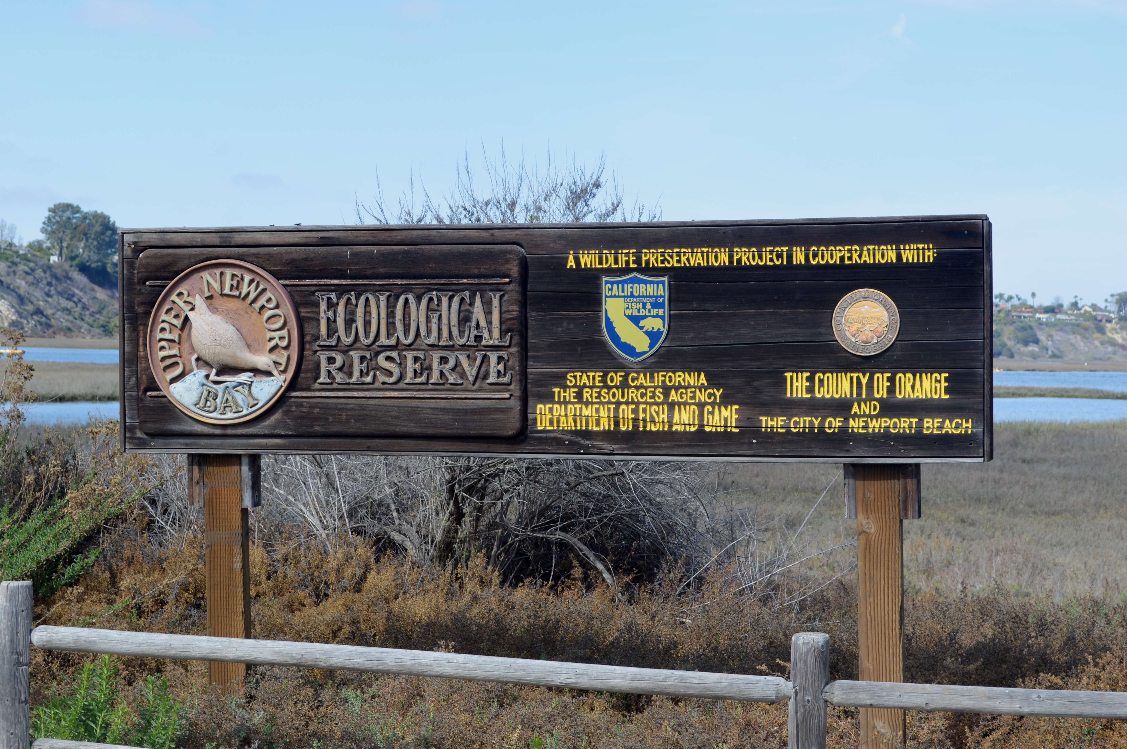 Wooden entrance sign for Upper Newport Bay Ecological Reserve with wetland and open water behind it.