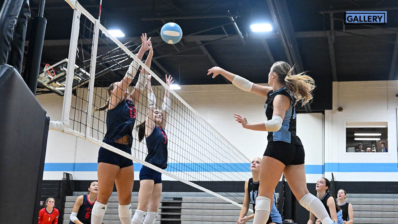 Volleyball players jump to block and spike the ball over the net in an indoor gymnasium.
