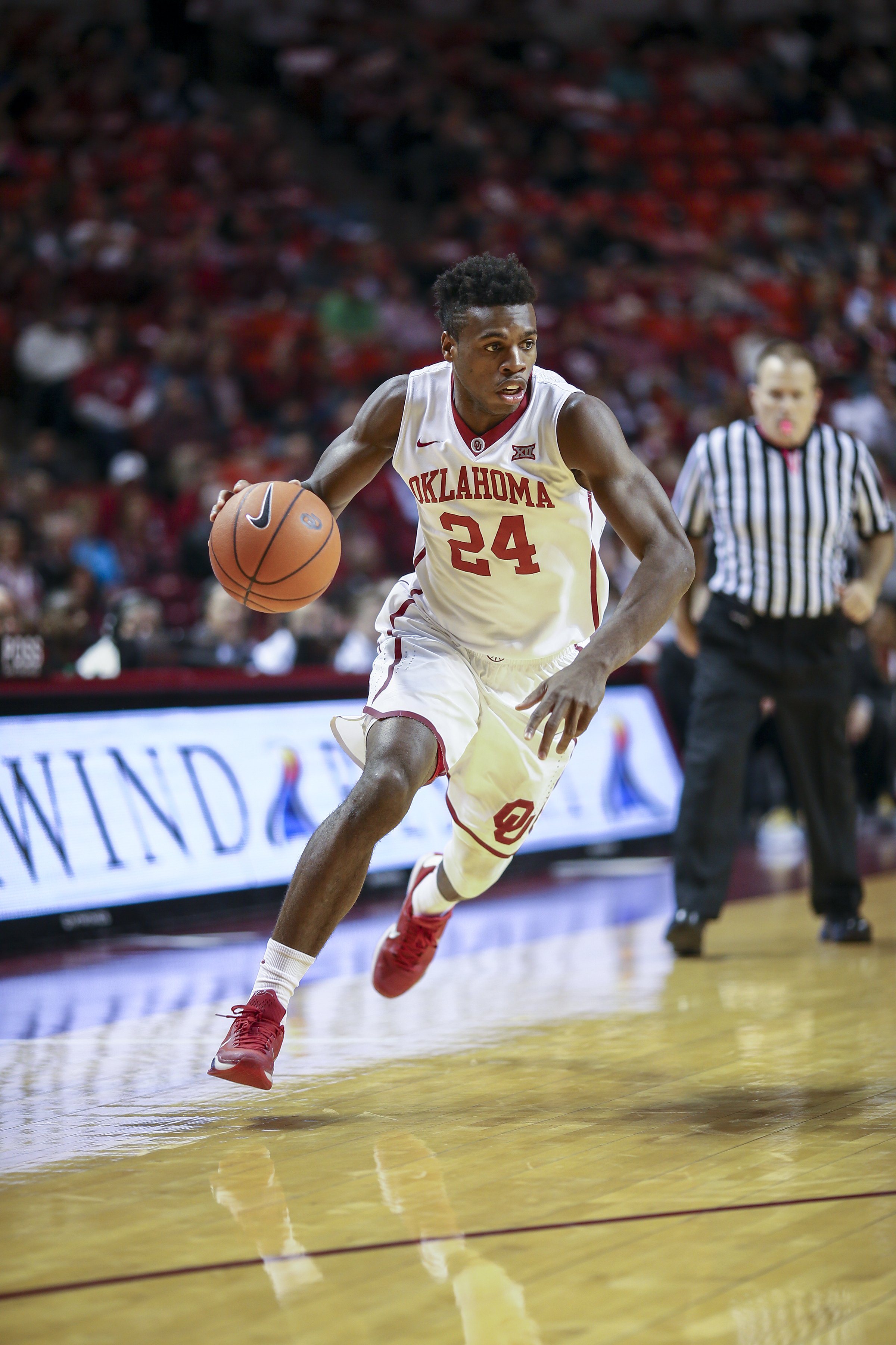 Basketball player in a red number 24 Sooners jersey dribbles on the court during a game.