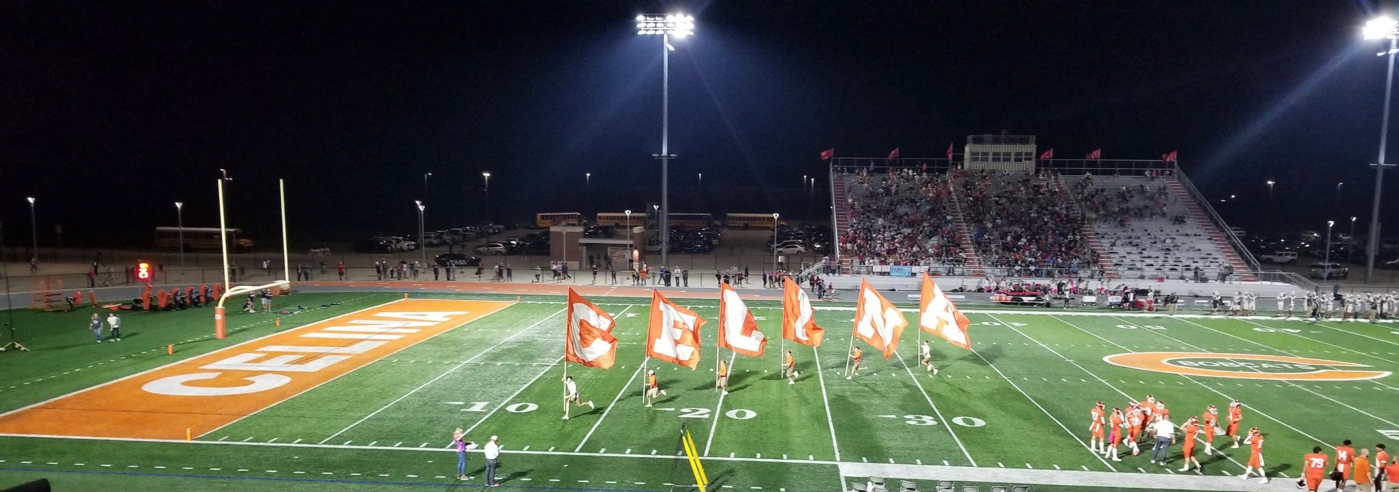 Football field at night under stadium lights with flags spelling out CELINA on the turf.