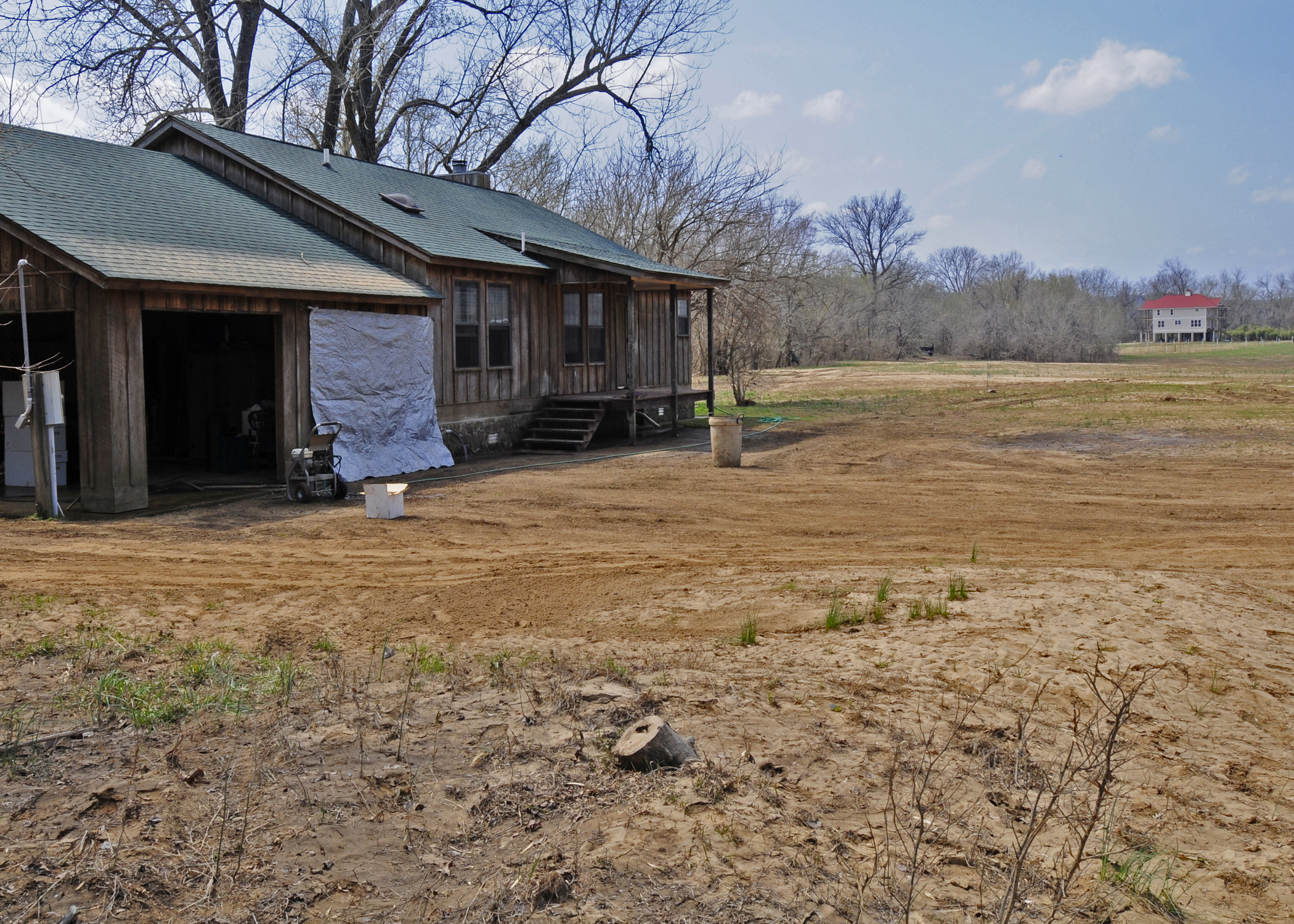 A flood-damaged ranch-style home with bare disturbed soil in the foreground and a blue tarp over a window.