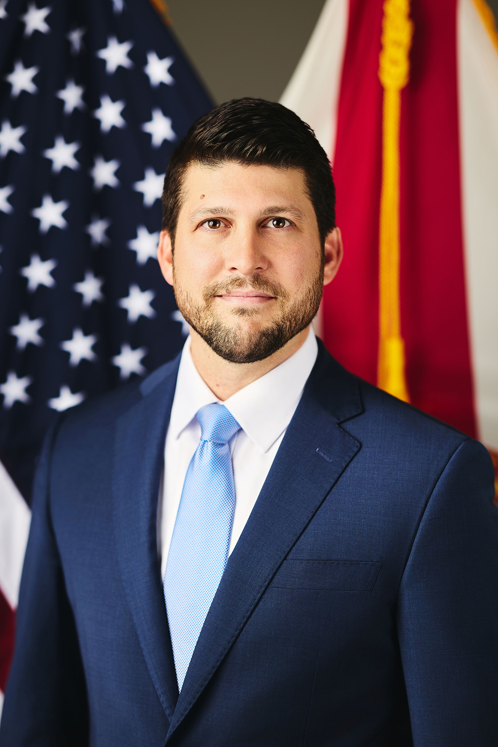 Man in navy suit and light blue tie poses before American and Florida state flags.