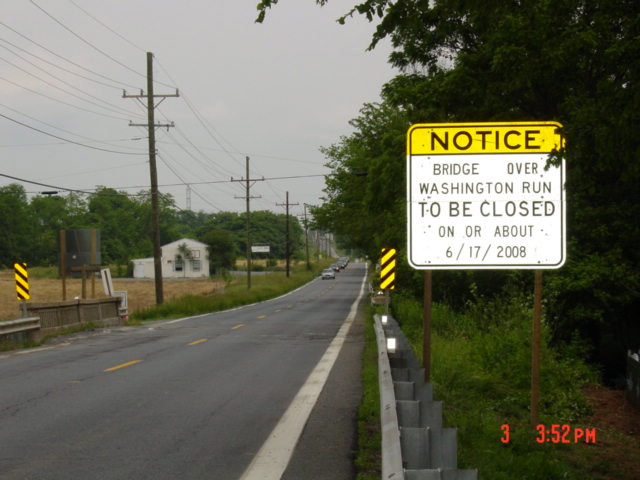 Yellow notice sign on a rural two-lane road warning of an upcoming bridge closure.