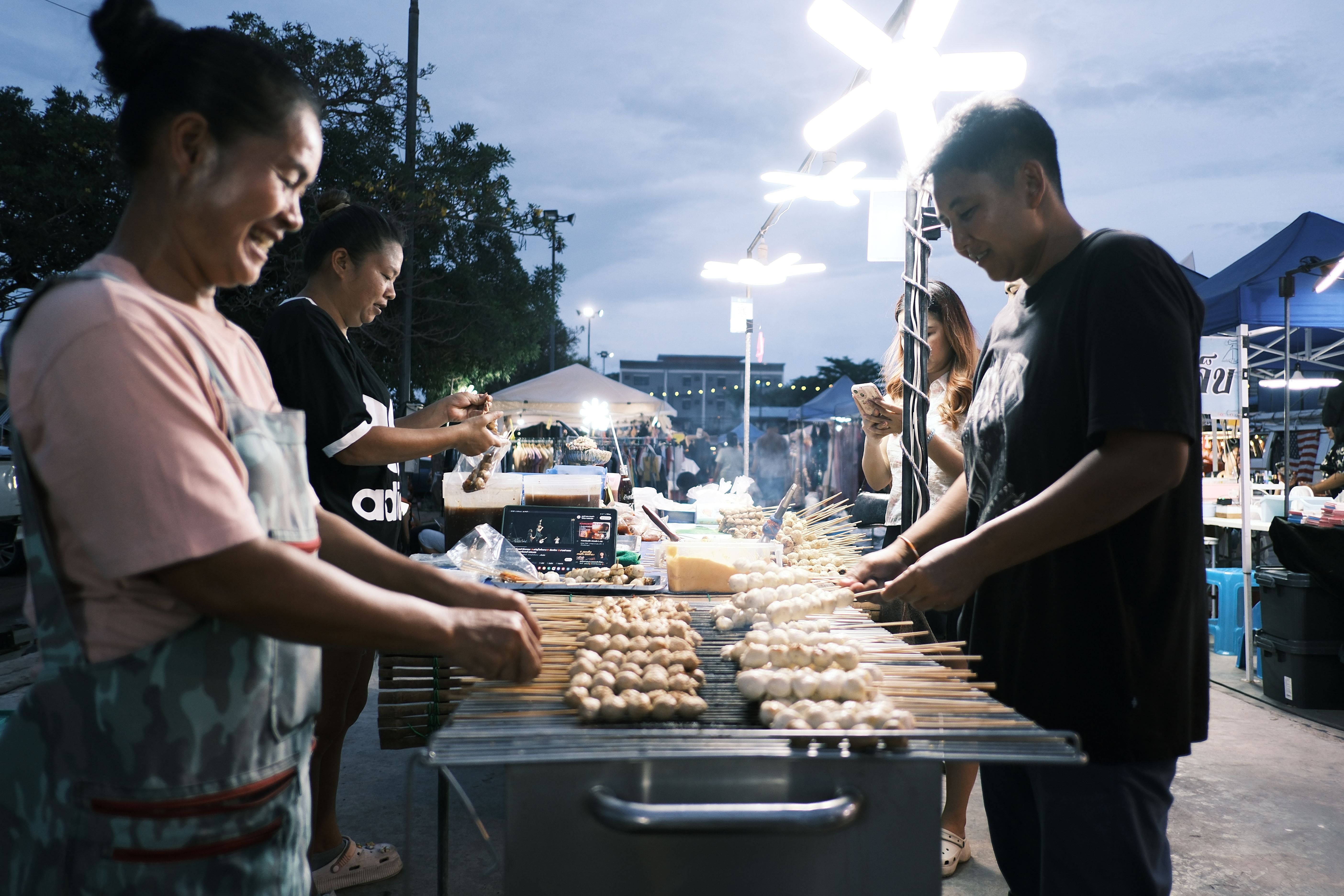 Vendors grilling rows of skewered food at a brightly lit outdoor market stall at dusk.