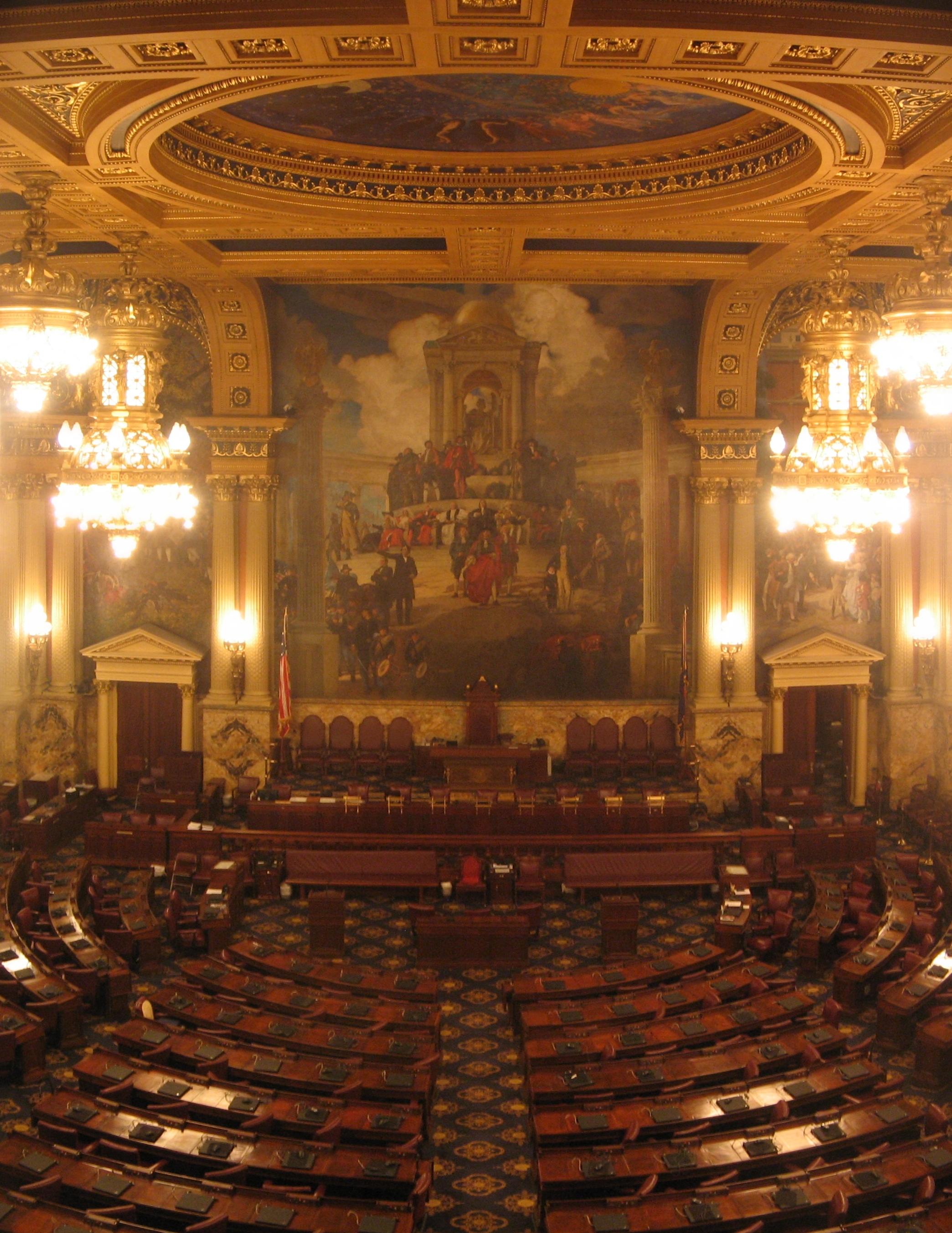 Ornate legislative chamber with rows of wooden desks, painted dome ceiling, and crystal chandeliers.