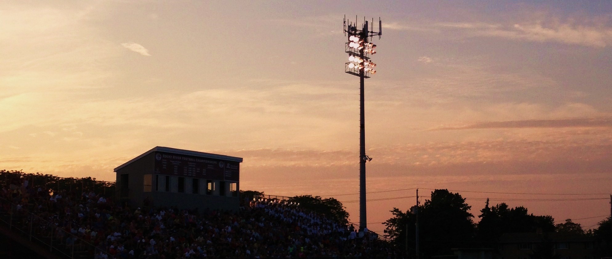 High school football stadium at sunset with a lit light tower and fans in the stands.