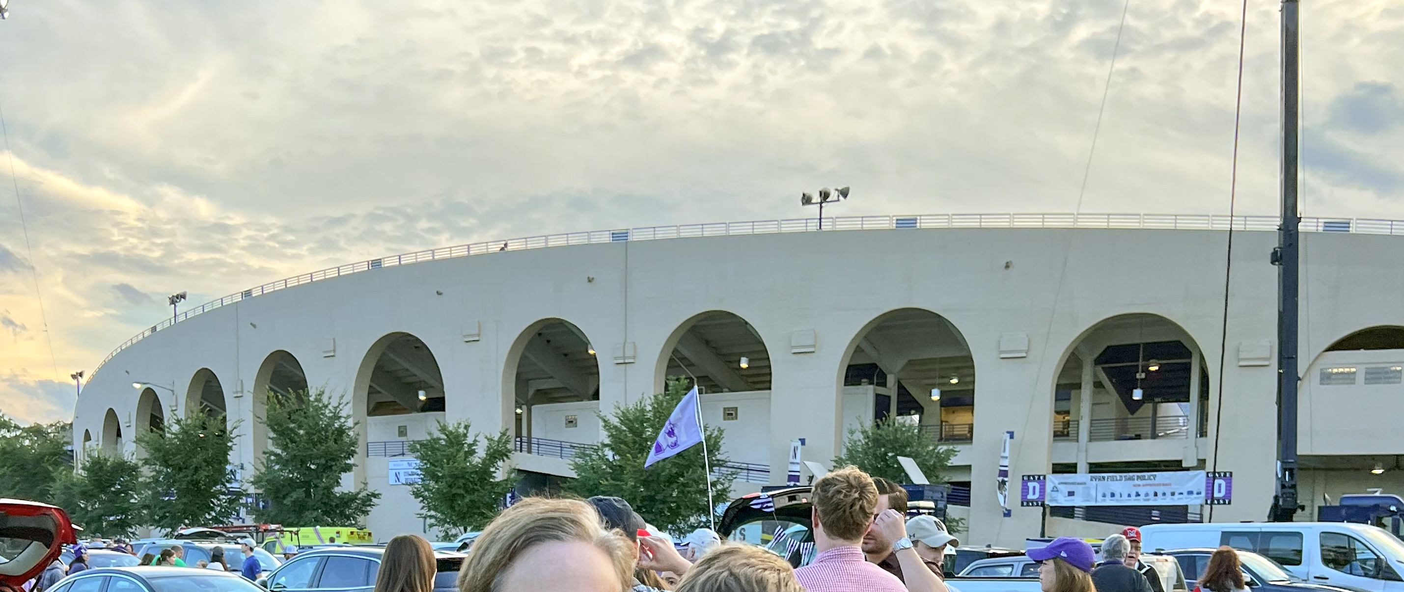Exterior of a large white stadium with arched openings, fans gathered in the parking lot at dusk.