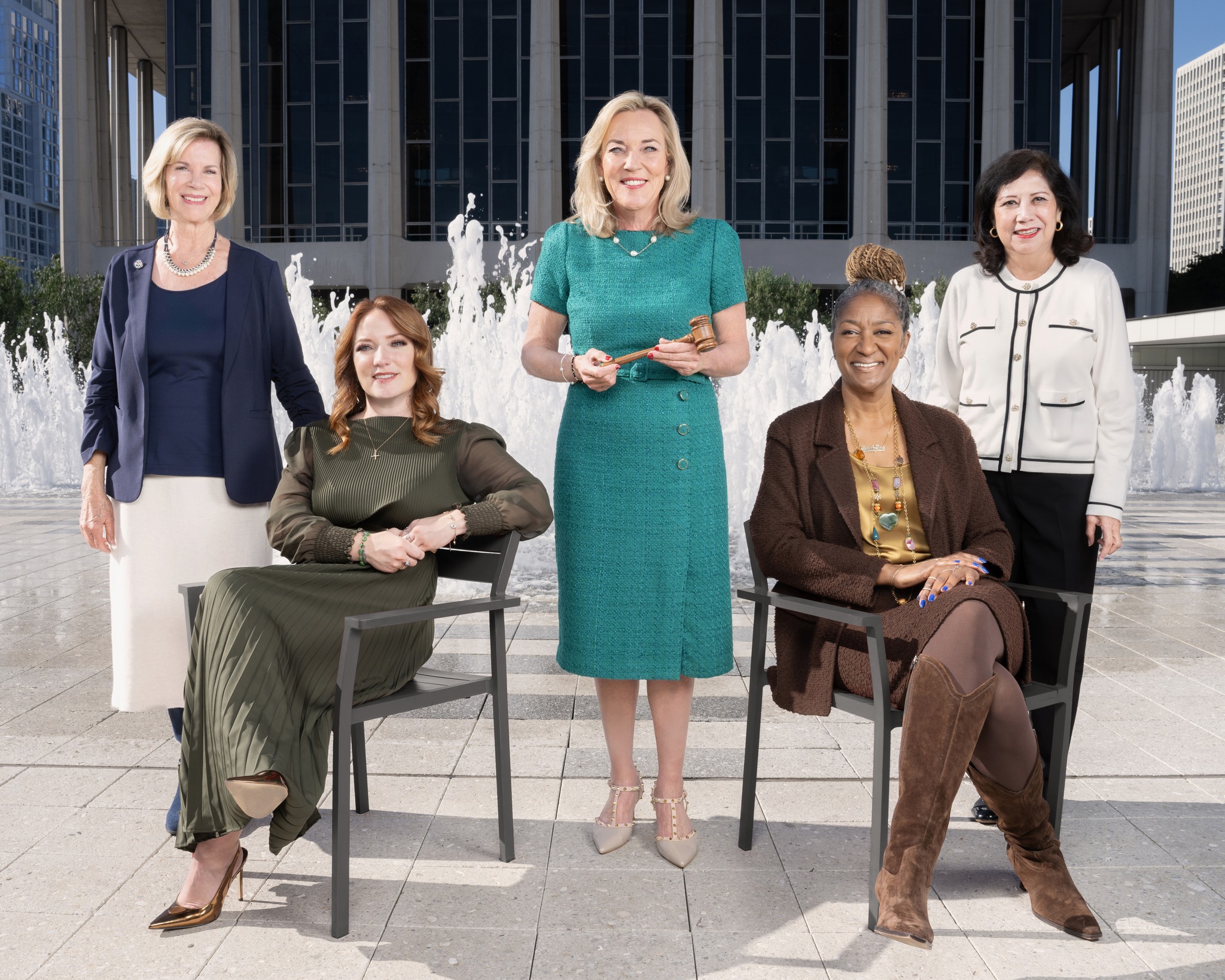 Five women posed outdoors in front of a fountain, three standing and two seated in chairs.