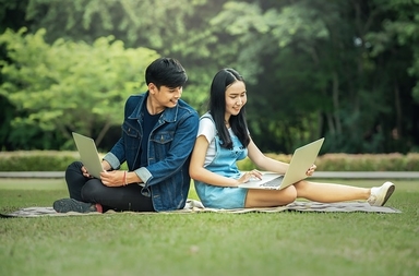 Two young people sit on grass outdoors, each working on a laptop computer.