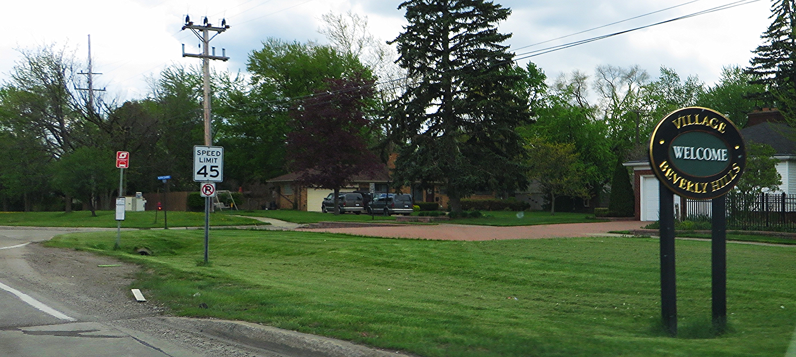 Circular village welcome sign for Beverly Hills, Michigan, along a tree-lined road.