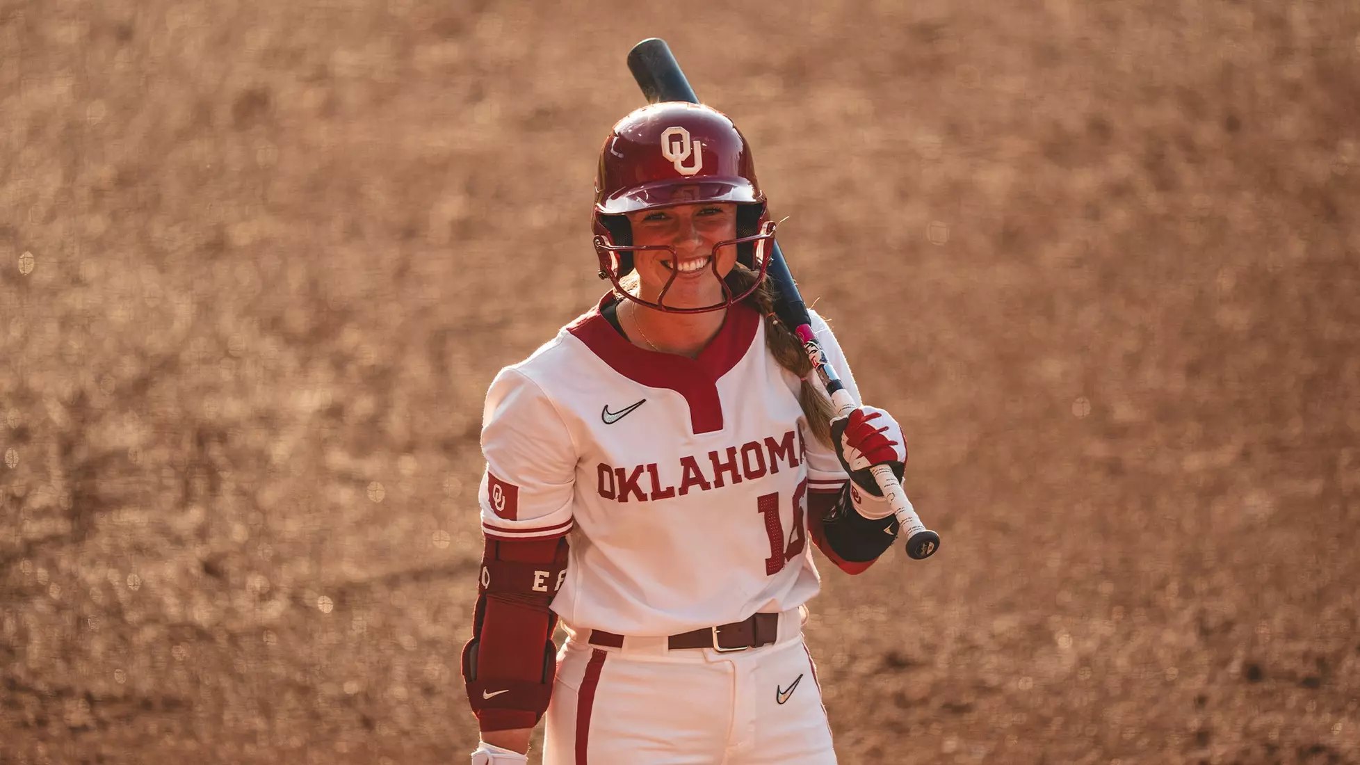 Softball player in white Oklahoma uniform smiling at bat on a dirt field.