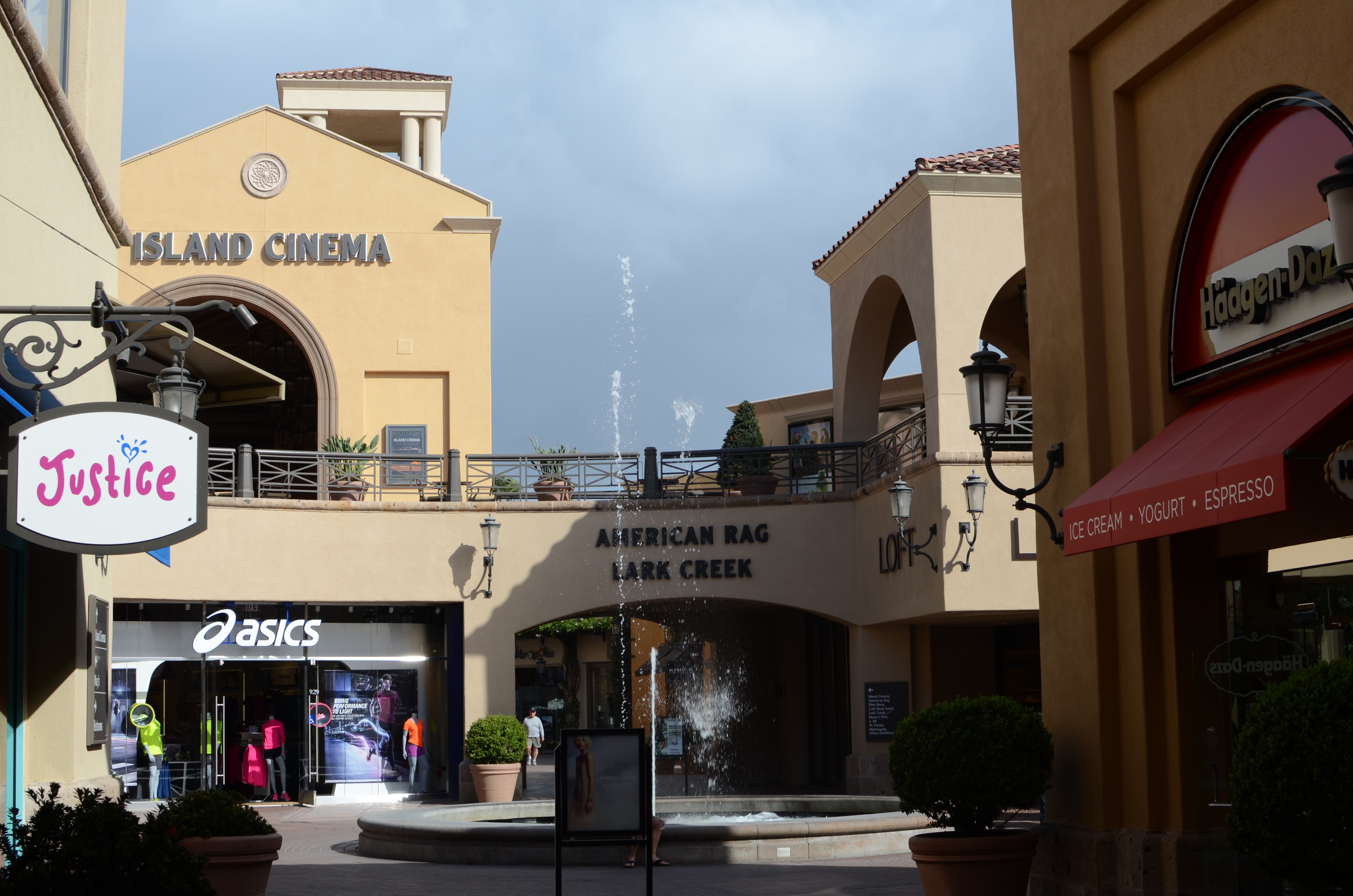 Outdoor shopping corridor with storefronts, a fountain, and palm trees at an open-air mall.