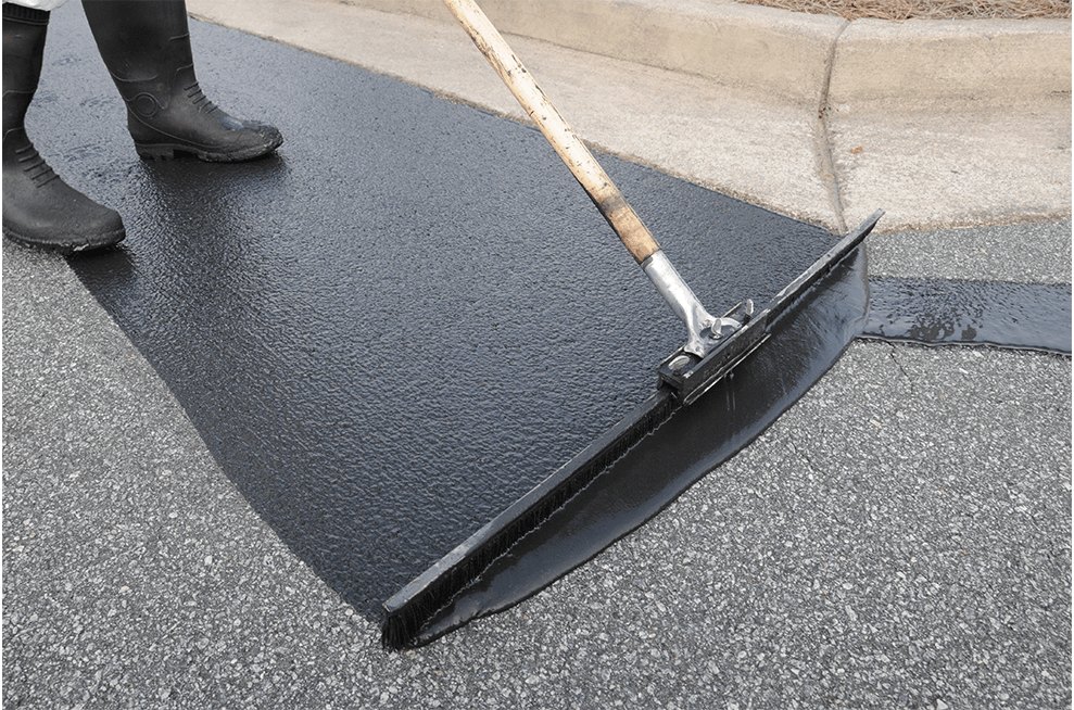 Worker in rubber boots using a long-handled squeegee to spread black sealant on asphalt.