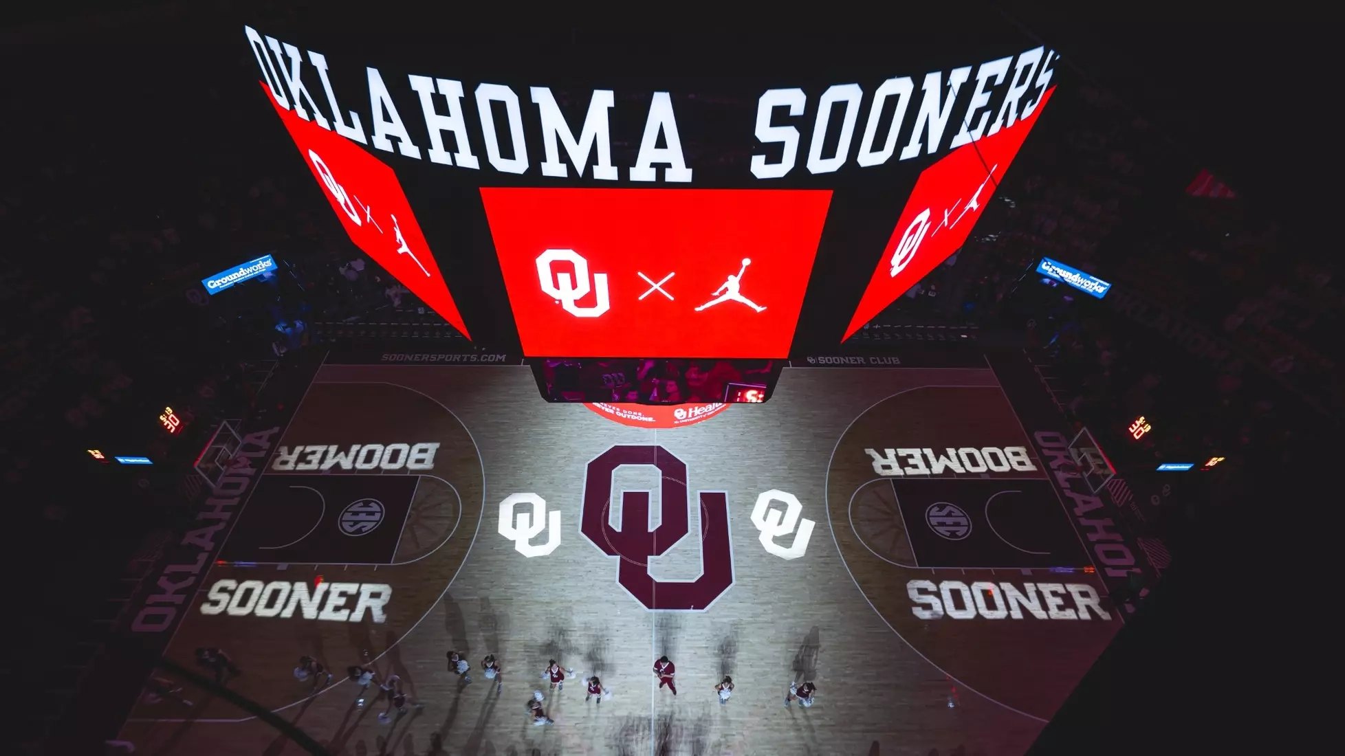 Aerial view of a basketball court with a large interlocking logo at center court and fans in the stands.
