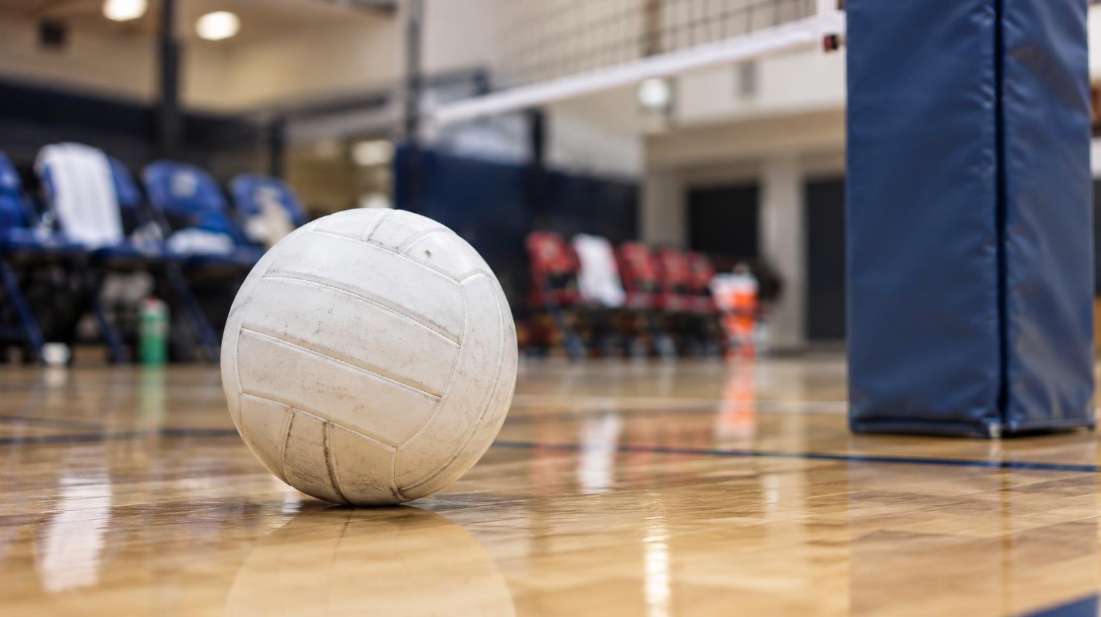 A white volleyball on a hardwood gym floor near a net post.