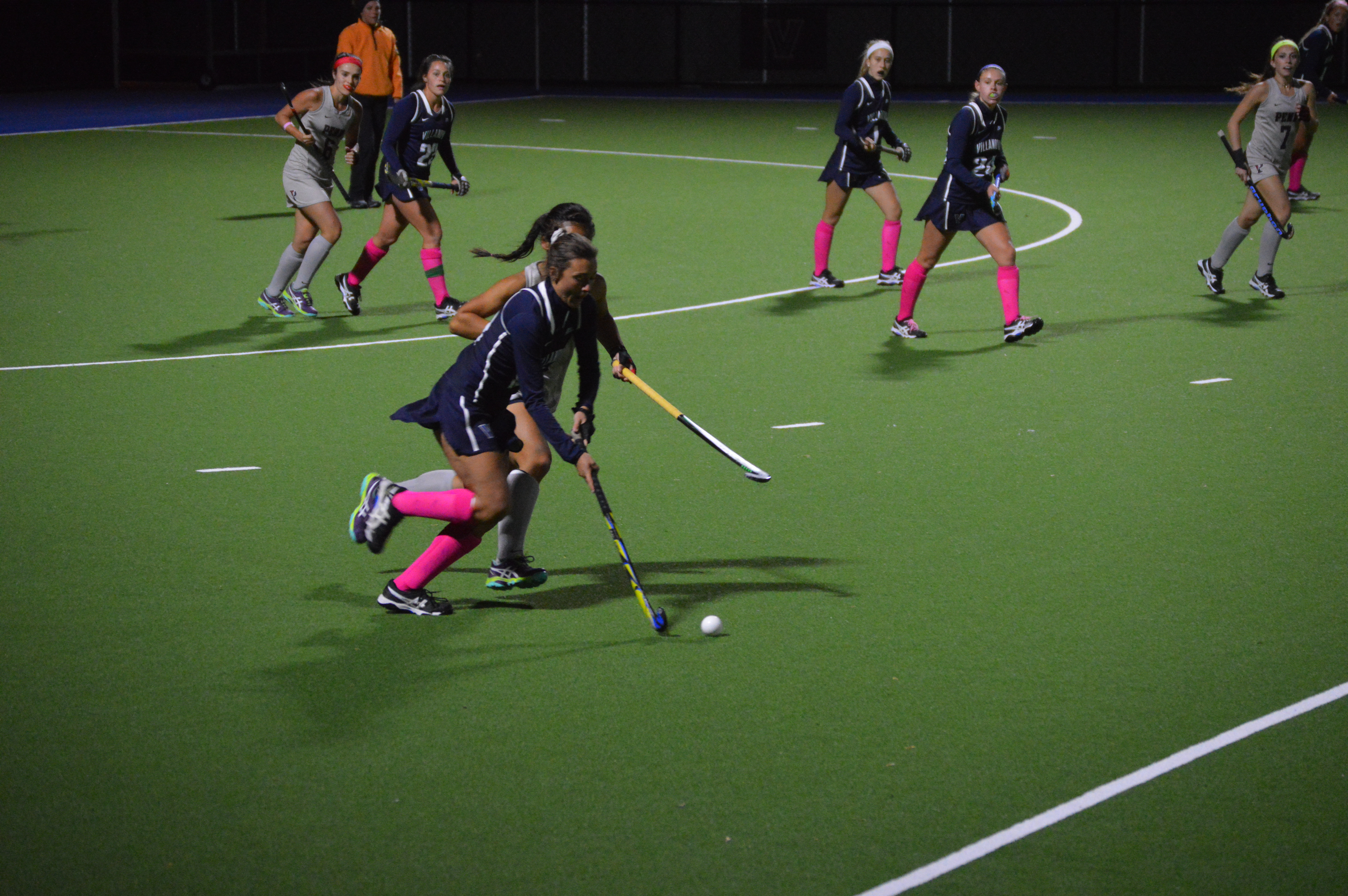 Female field hockey players in dark uniforms competing on a lit turf field at night.