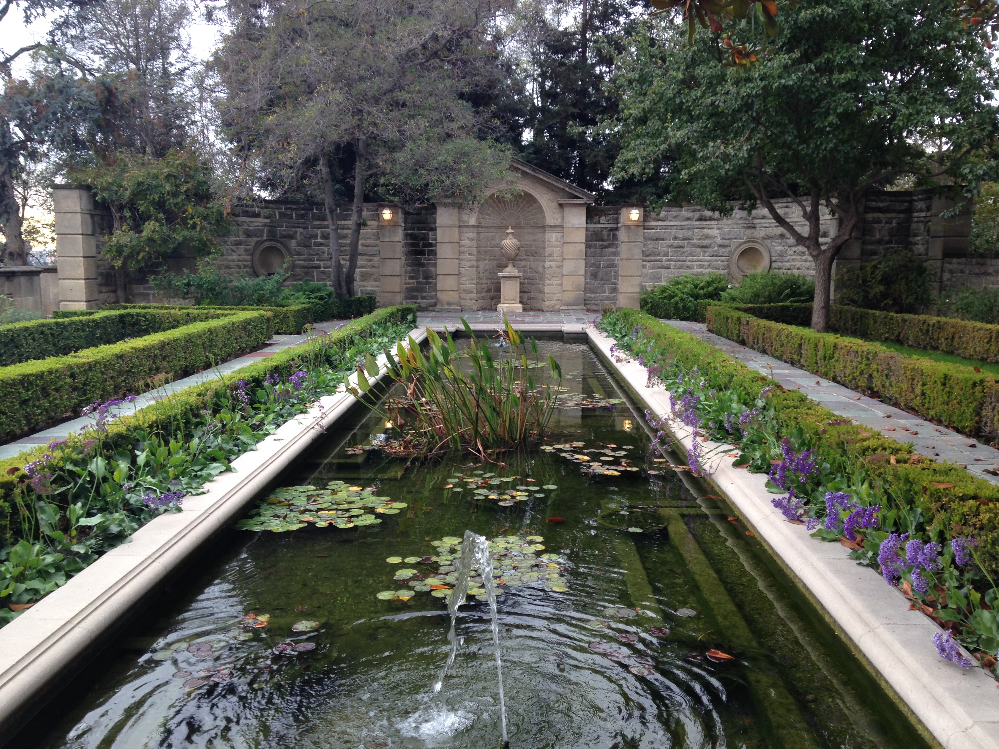 Rectangular reflecting pool with lily pads and a small fountain, flanked by hedges and a stone wall.